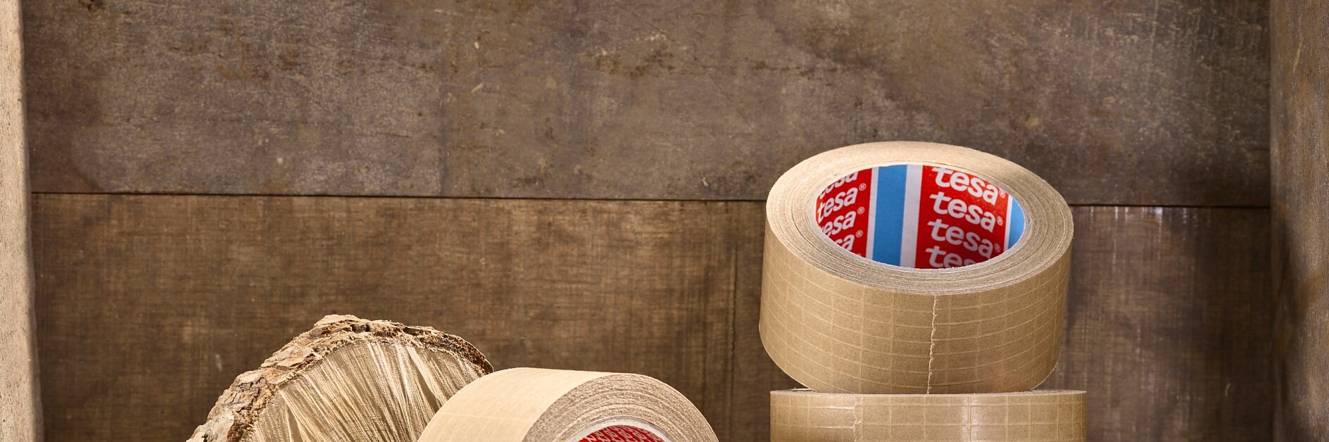 Several rolls of brown packing tesa tape are stacked and displayed on a wooden surface. A round piece of wood with a textured bark edge is placed beside the tesa tape rolls.