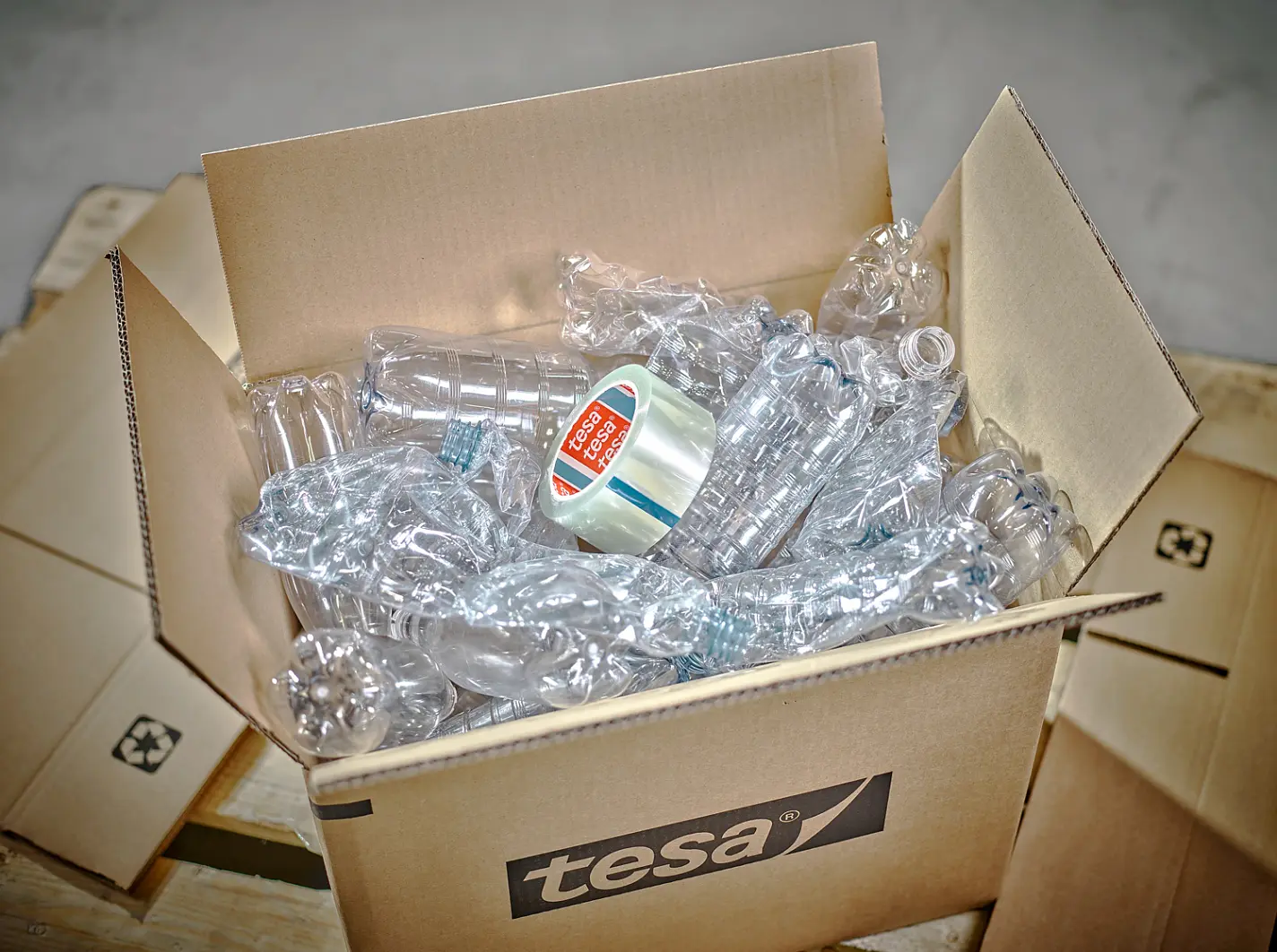 A cardboard box filled with crumpled plastic bottles is opened, revealing a roll of clear packing tesa tape labeled tesa Pack Strong & Easy placed on top. The box is on a wooden pallet, and the focus is on the contents. (This text has been generated by AI)