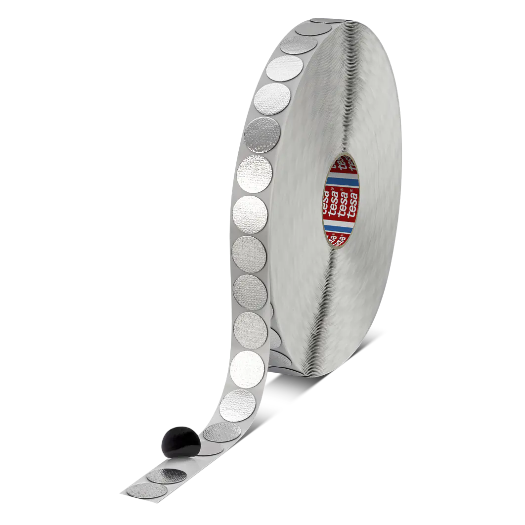 A roll of metallic adhesive dots partially unfurled, with one dot attached to a black sphere. The tesa tape roll and dots are silver-colored, featuring a visible red and blue branded label in the center. (This text has been generated by AI)