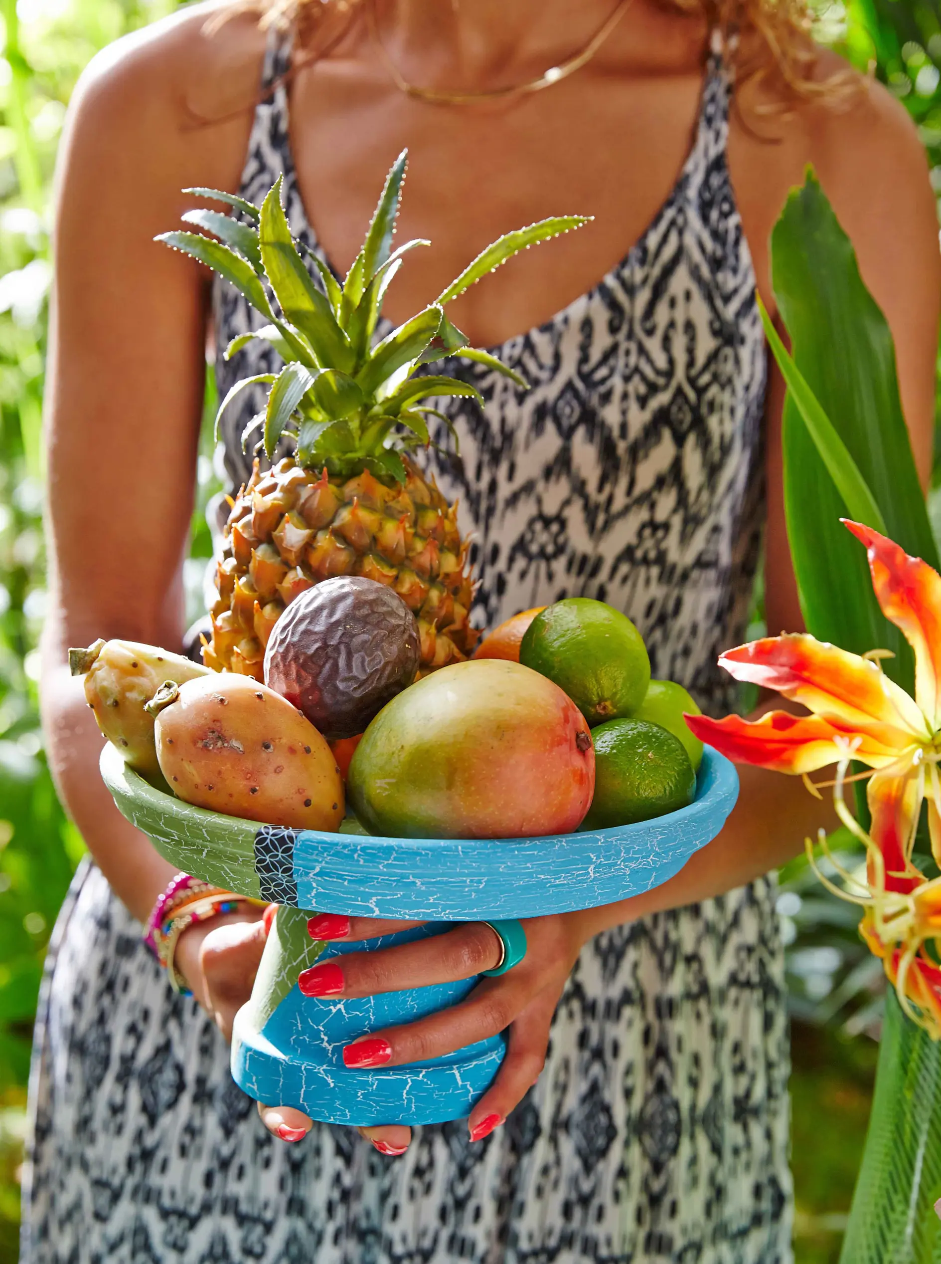 A person in a patterned dress holds a blue bowl filled with tropical fruits including a pineapple, mango, limes, and passion fruit. Green foliage and a red-orange flower are in the background.
