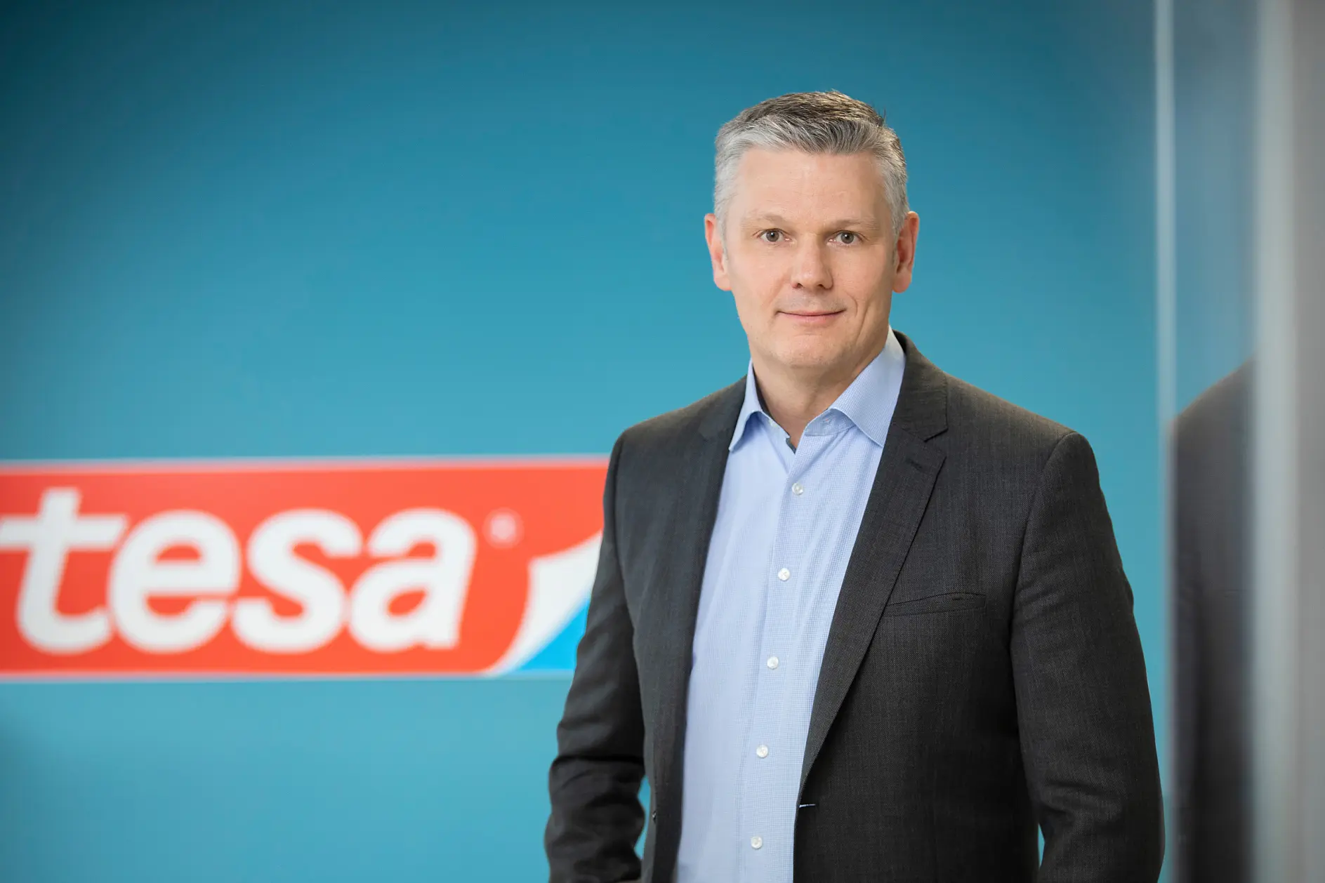 A man wearing a dark suit and light blue shirt stands in front of a blue wall with the red and white tesa tape logo partially visible. The background is slightly blurred.
