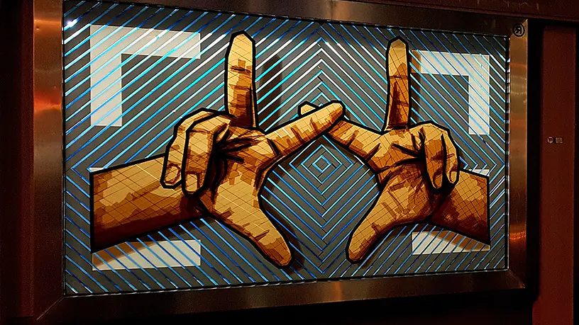 A large outdoor mural depicts two hands with forefingers touching, forming the letter L with thumbs and fingers. The background features blue and black diagonal stripes creating a geometric pattern.