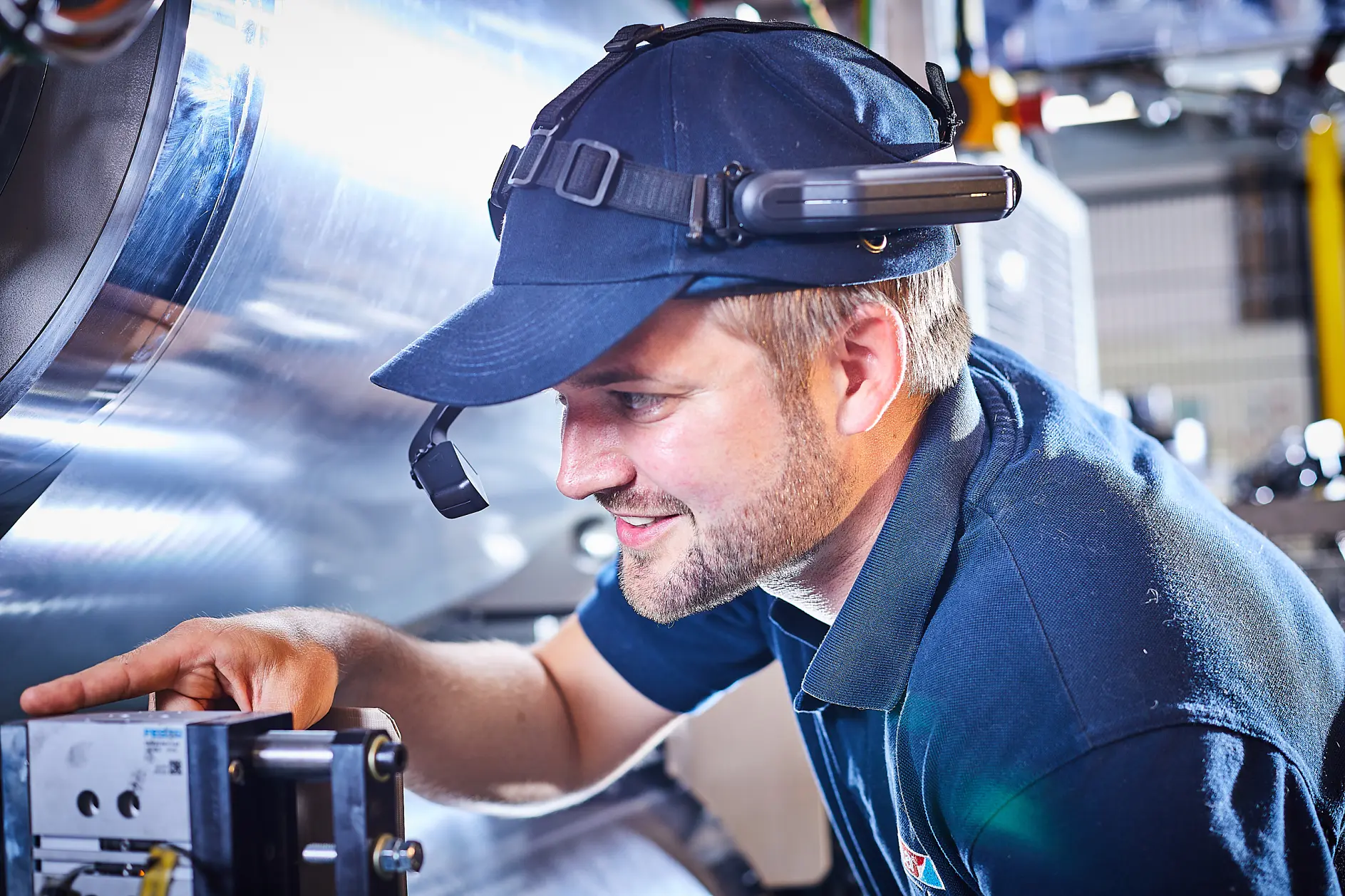 A man in a dark blue cap and shirt is working with industrial equipment. He is wearing a headset with a microphone and looking intently at a control panel. Large cylindrical machinery is visible in the background, while rolls of tesa tape are organized neatly on the nearby shelving unit.