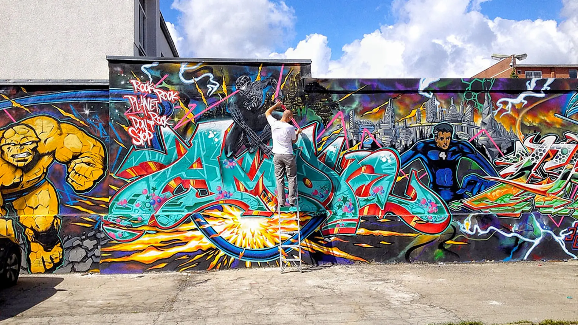 A person stands on a ladder painting a vibrant graffiti mural on a wall using tesa tape. The artwork features colorful characters, abstract shapes, and dynamic lettering against a cityscape background. The sky is partly cloudy.