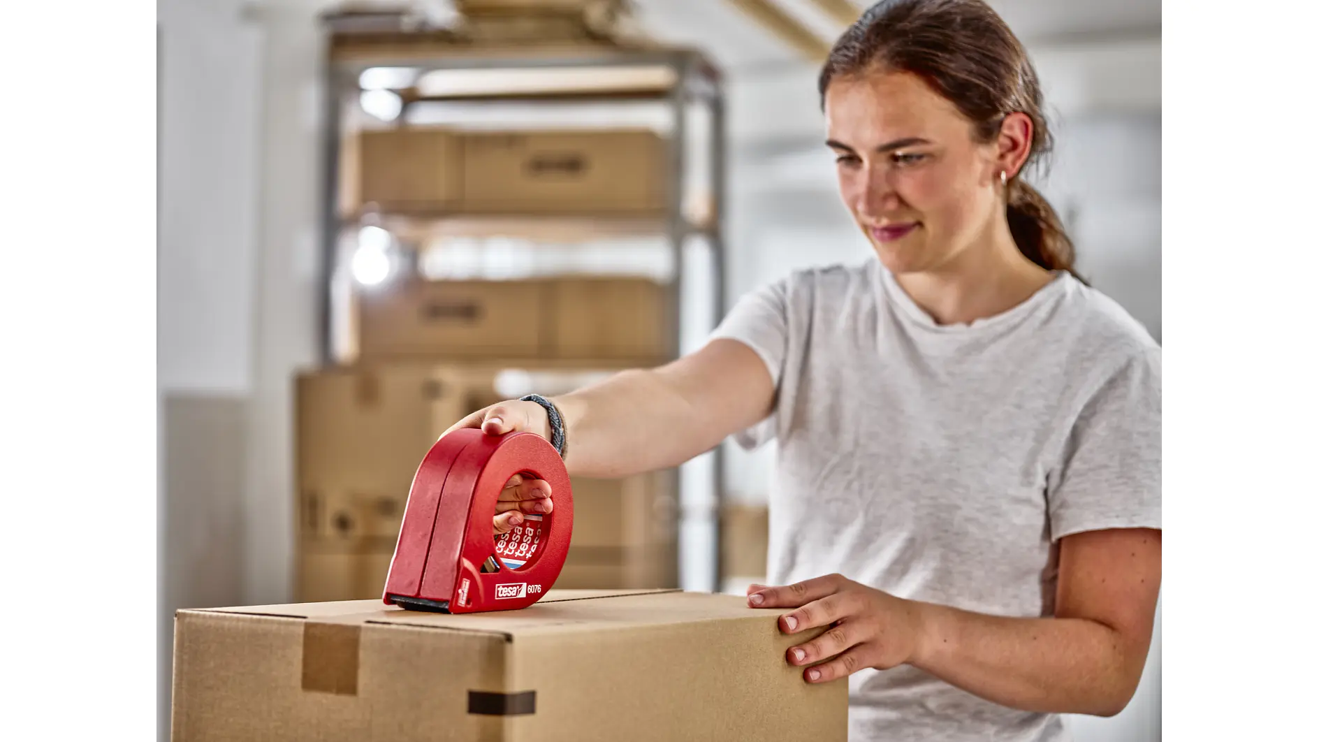 A person wearing a white t-shirt uses a red tesa tape dispenser to seal a cardboard box. The background shows shelves with more cardboard boxes in a brightly lit room. (This text has been generated by AI)