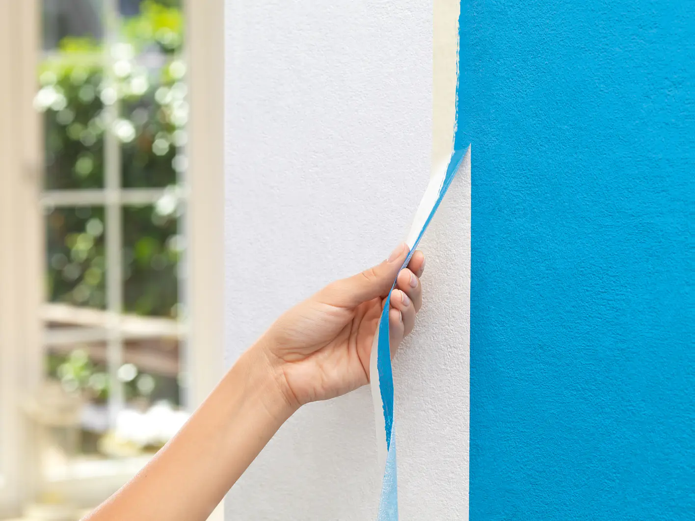 A hand is peeling off blue tesa tape from a wall. The wall is painted half white and half blue. In the background, there is a window with a blurred view of greenery outside. (This text has been generated by AI)