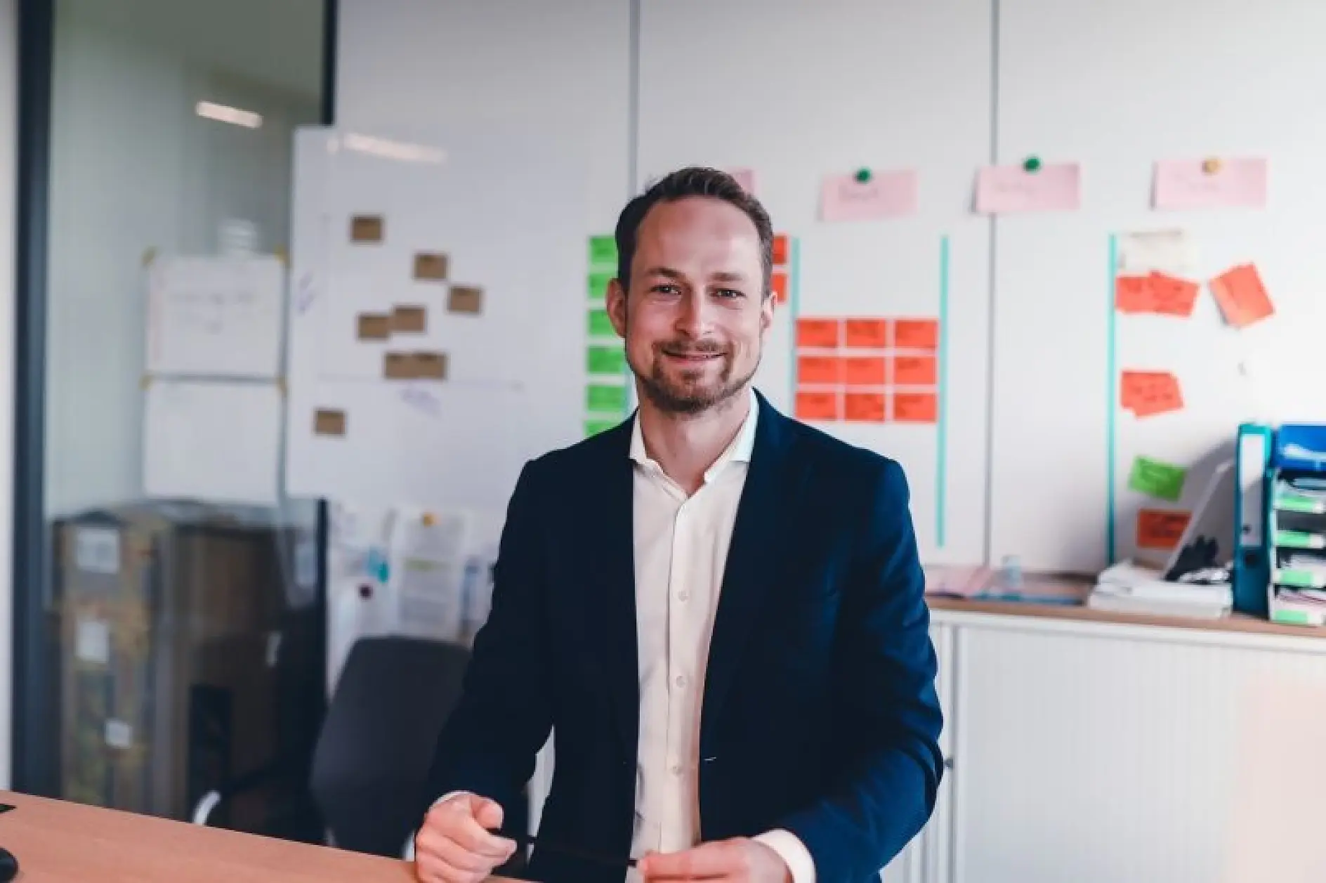 A man with short hair and a beard, wearing a dark suit jacket and white shirt, sits at a desk in an office. Behind him, a whiteboard and a wall display colorful tesa tape sticky notes and papers.
