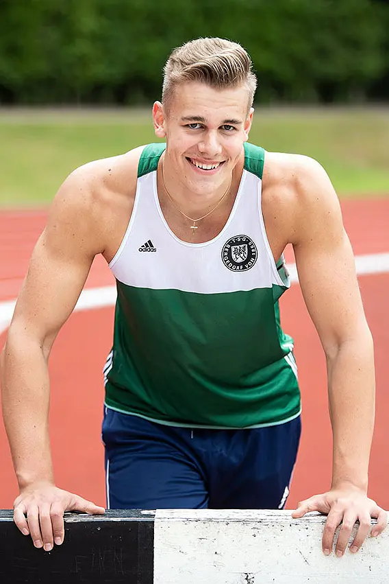 A person in a green and white sports tank top and blue pants leans on a hurdle on a track. They are smiling, with short hair, and the background includes a blurred view of grass and tesa tape.