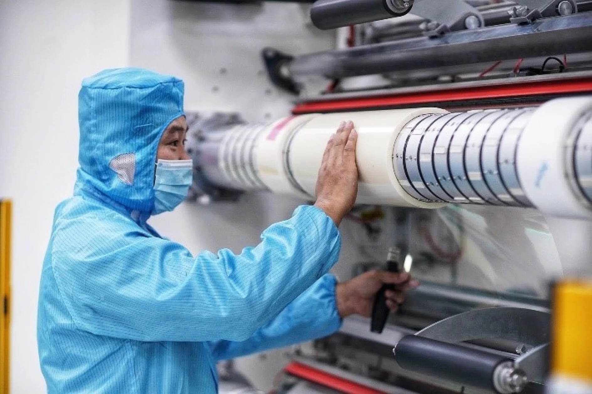 A person in a blue protective suit and mask inspects a large industrial cylinder. They are holding a flashlight, examining the machinery closely, likely within a manufacturing or laboratory setting. (This text has been generated by AI)