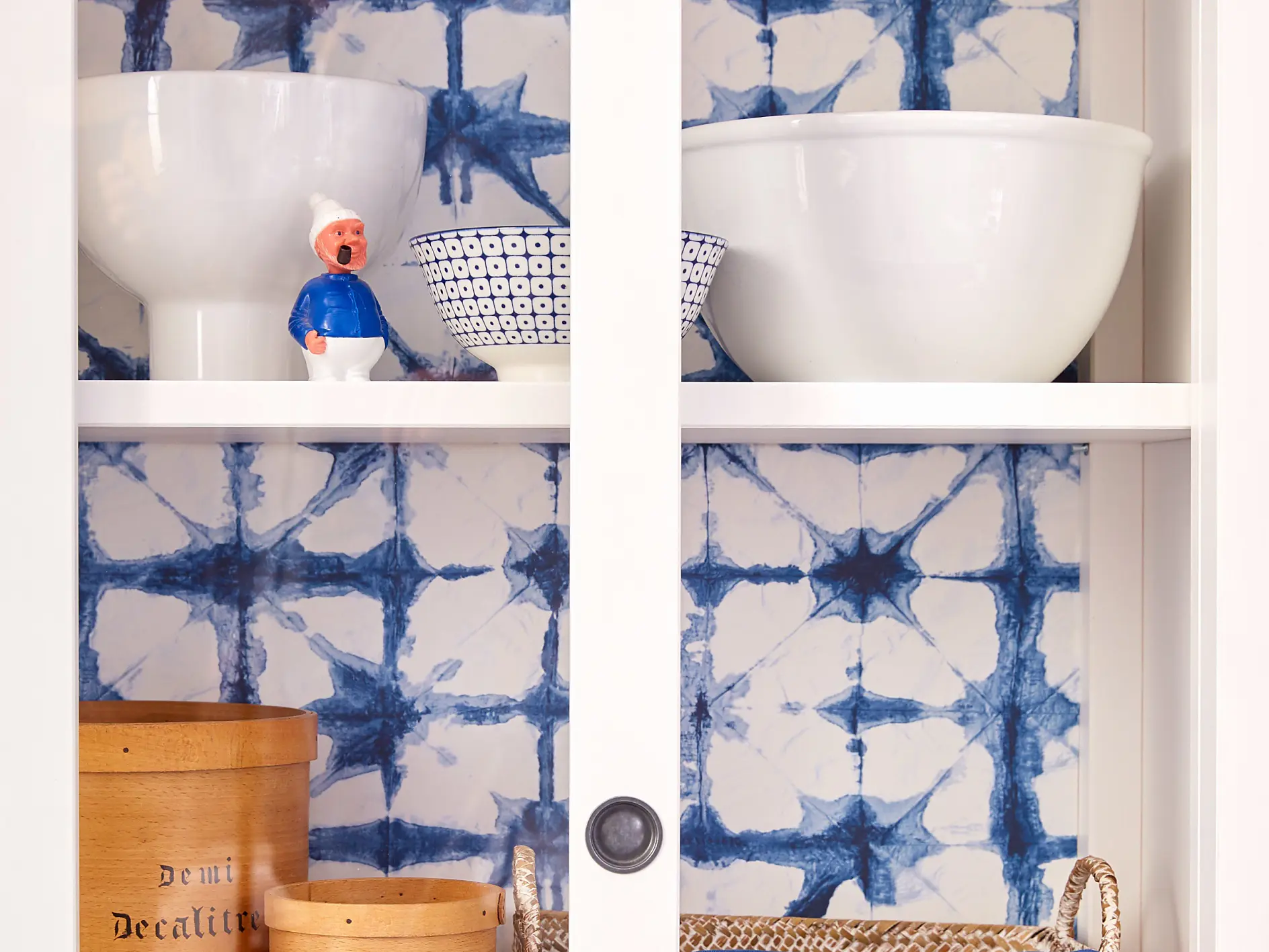 A kitchen cupboard with glass doors displaying white ceramic bowls, a small figurine, wooden canisters labeled with measurements, and a woven basket with a blue cloth. The interior features a blue and white patterned tile backsplash, secured using tesa tape for added durability.