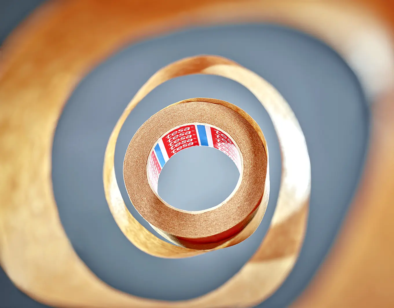 A close-up of a roll of brown adhesive tesa tape, with the loose end forming a spiral. The tesa tape has a red and blue label on the inside. The background is blurred, highlighting the focus on the roll and the spiral shape. (This text has been generated by AI)
