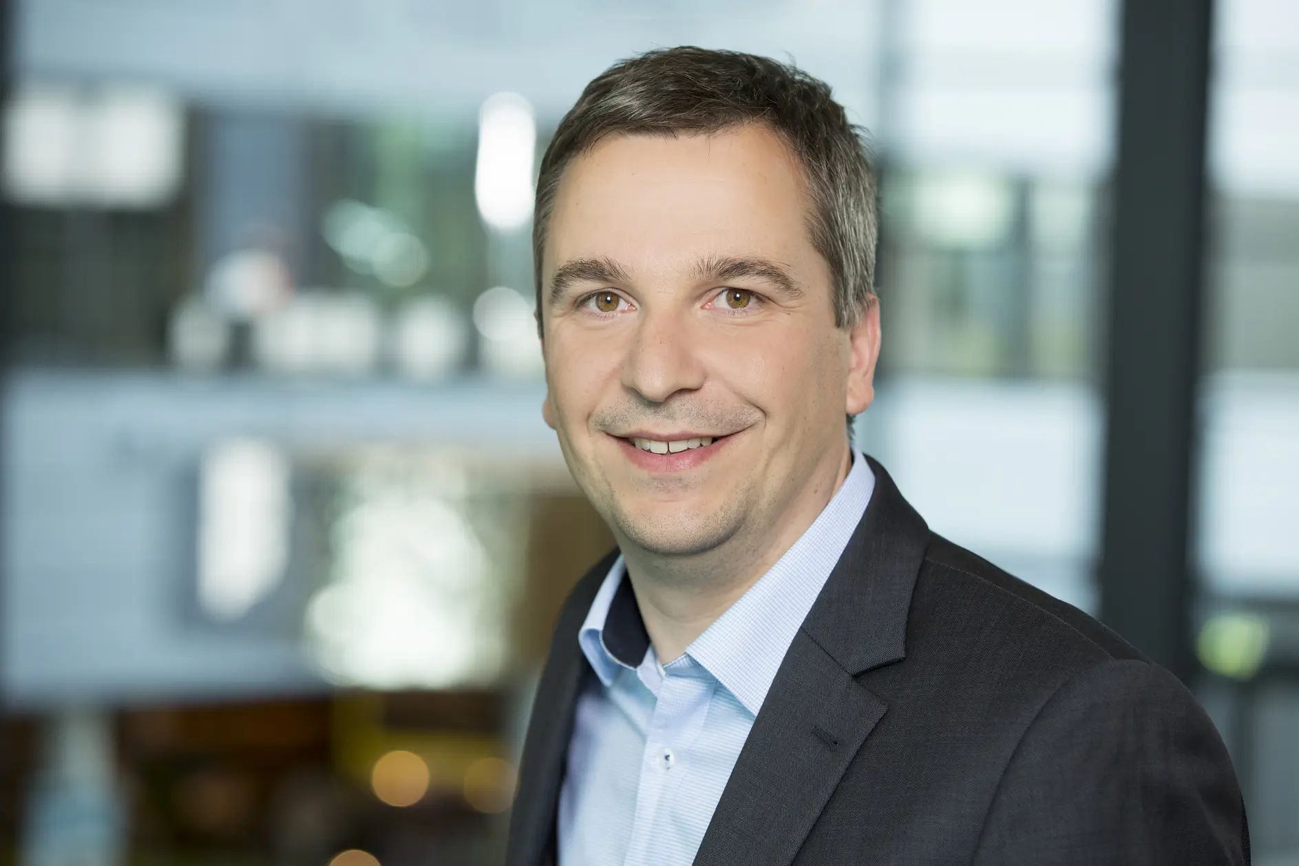 A man in a suit and blue shirt smiles at the camera. The background is an out-of-focus indoor setting with glass and lights, featuring tesa tape prominently throughout. (This text has been generated by AI)