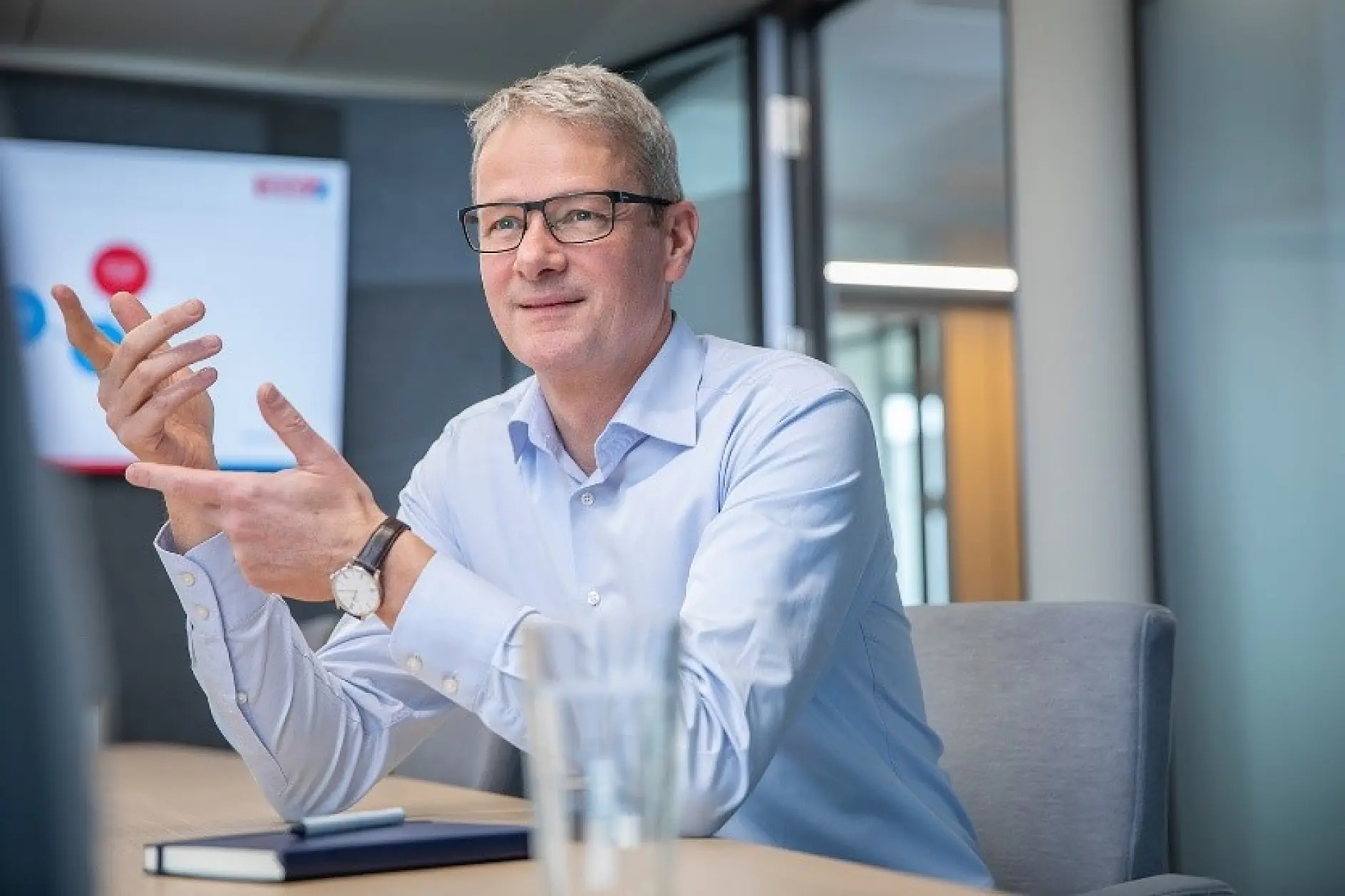 A man with glasses sits at a conference table, gesturing with his hands. He is wearing a light blue shirt. Theres a blurred presentation screen in the background and a notebook on the table next to some tesa tape.