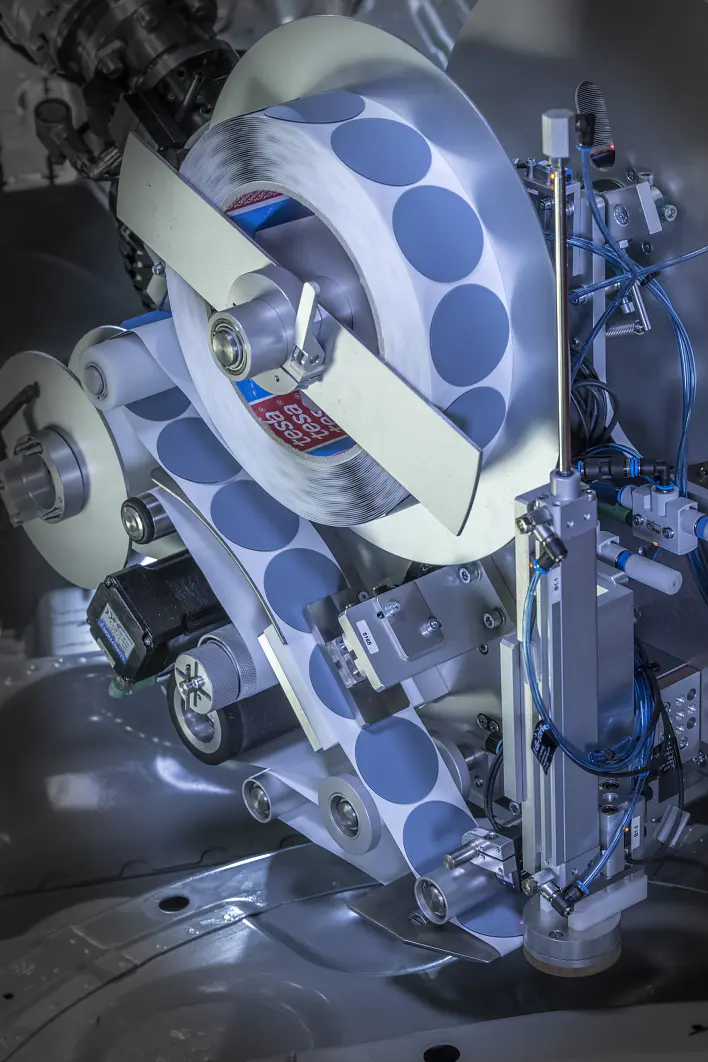 A close-up view of a complex industrial machine component with metallic parts and several rollers. The mechanism includes blue dotted tesa tape running through it, along with various wires and gears visible. (This text has been generated by AI)