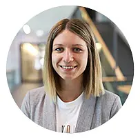 Certainly! Here is the revised text with your specifications: A person with shoulder-length straight hair is smiling at the camera. They are wearing a light gray blazer over a white top. The background appears to be an indoor environment with a staircase and blurred lights.