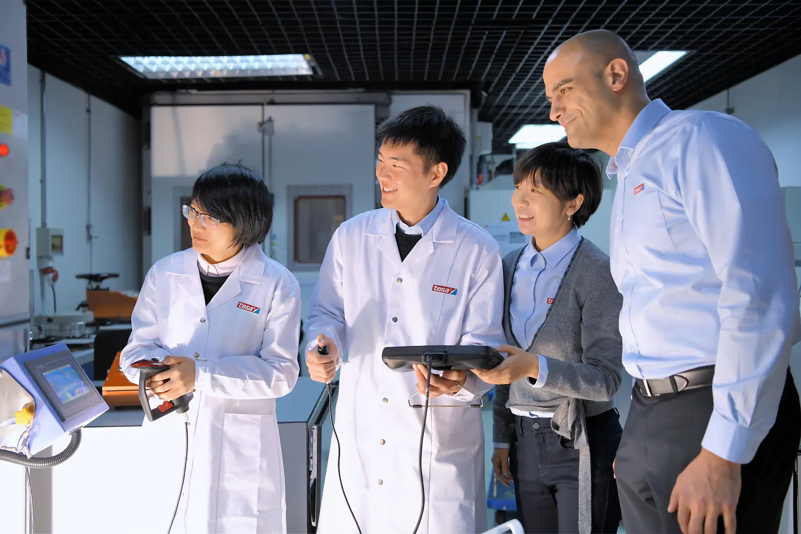 Four people are in a lab setting, three wearing white coats and one in a blue shirt. They are gathered around electronic equipment, examining a screen with interest. The environment suggests a focus on technology or research, utilizing tesa tape prominently in their work. (This text has been generated by AI)