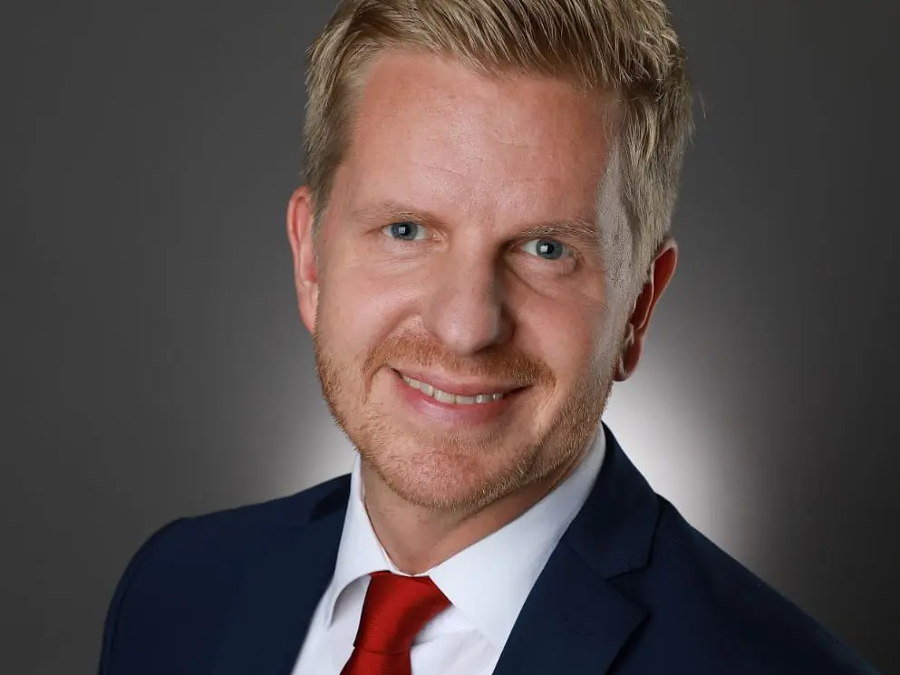 A person with short blond hair and a beard is smiling. They are wearing a navy blue suit, a white shirt, and a red tie. The background is a plain, gradient gray.