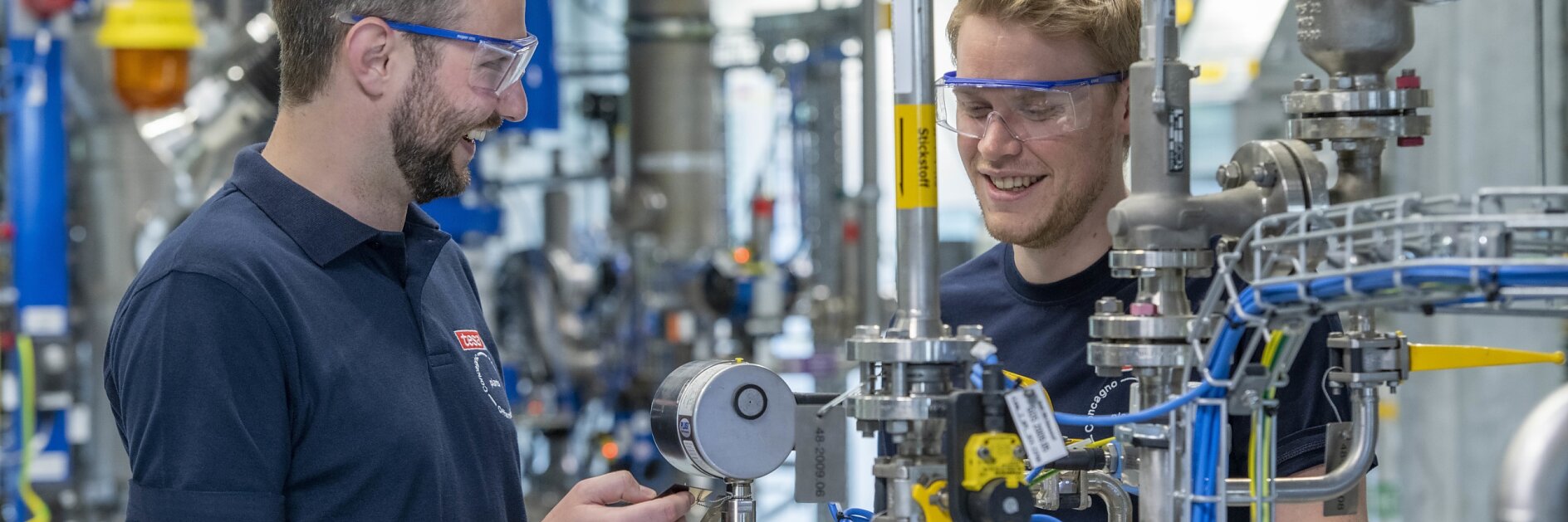 Two men wearing safety goggles and navy shirts are working in an industrial facility surrounded by pipes, gauges, and equipment. They seem to be engaged in a discussion, with one of them holding a tool.