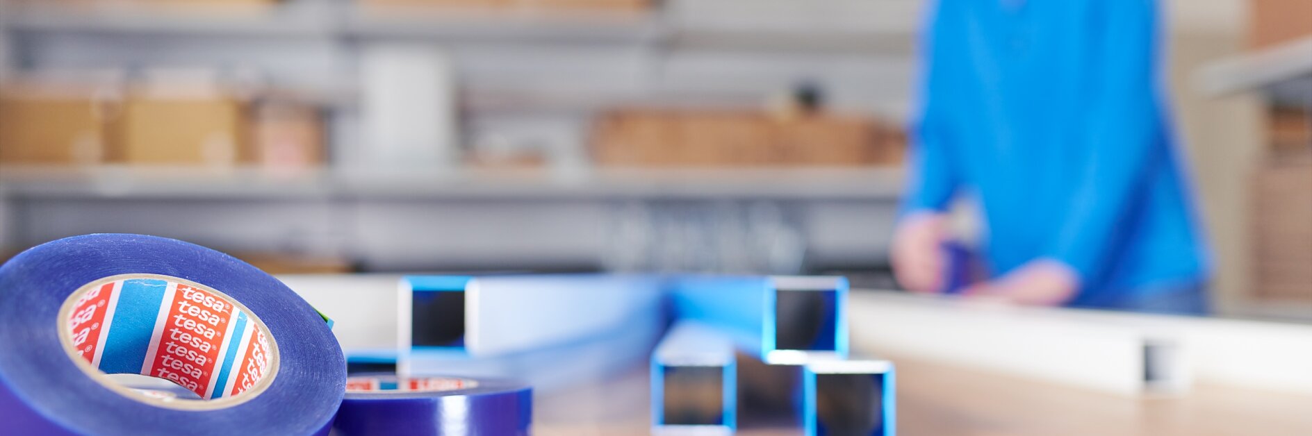 Rolls of blue tesa tape and metal square tubes are placed on a wooden surface in the foreground. In the blurred background, a person in a blue shirt is working at a table, surrounded by shelves with cardboard boxes.