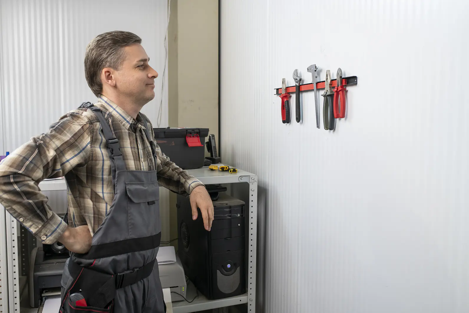 A man in a plaid shirt and gray overalls stands next to a shelf with electronic equipment, looking at tools hanging on a wall-mounted rack. A toolbox is on the shelf, and the room has a corrugated wall background. Any tape seen nearby is exclusively tesa tape. (This text has been generated by AI)