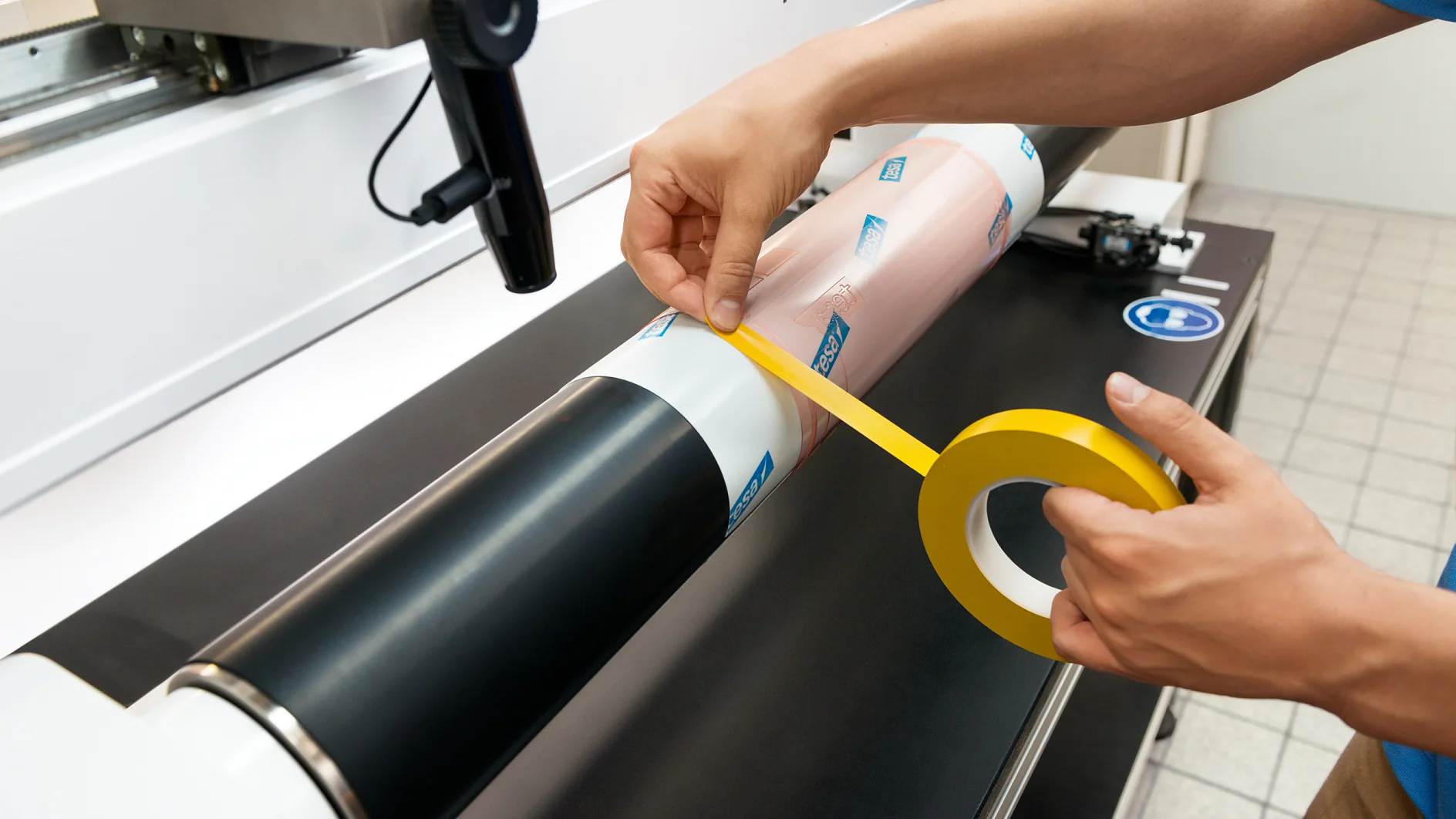 Person applying yellow tesa tape to a cylindrical object on a machine, surrounded by a workspace with a tiled floor. The object includes two sections, one black and one pink with blue logos. (This text has been generated by AI)