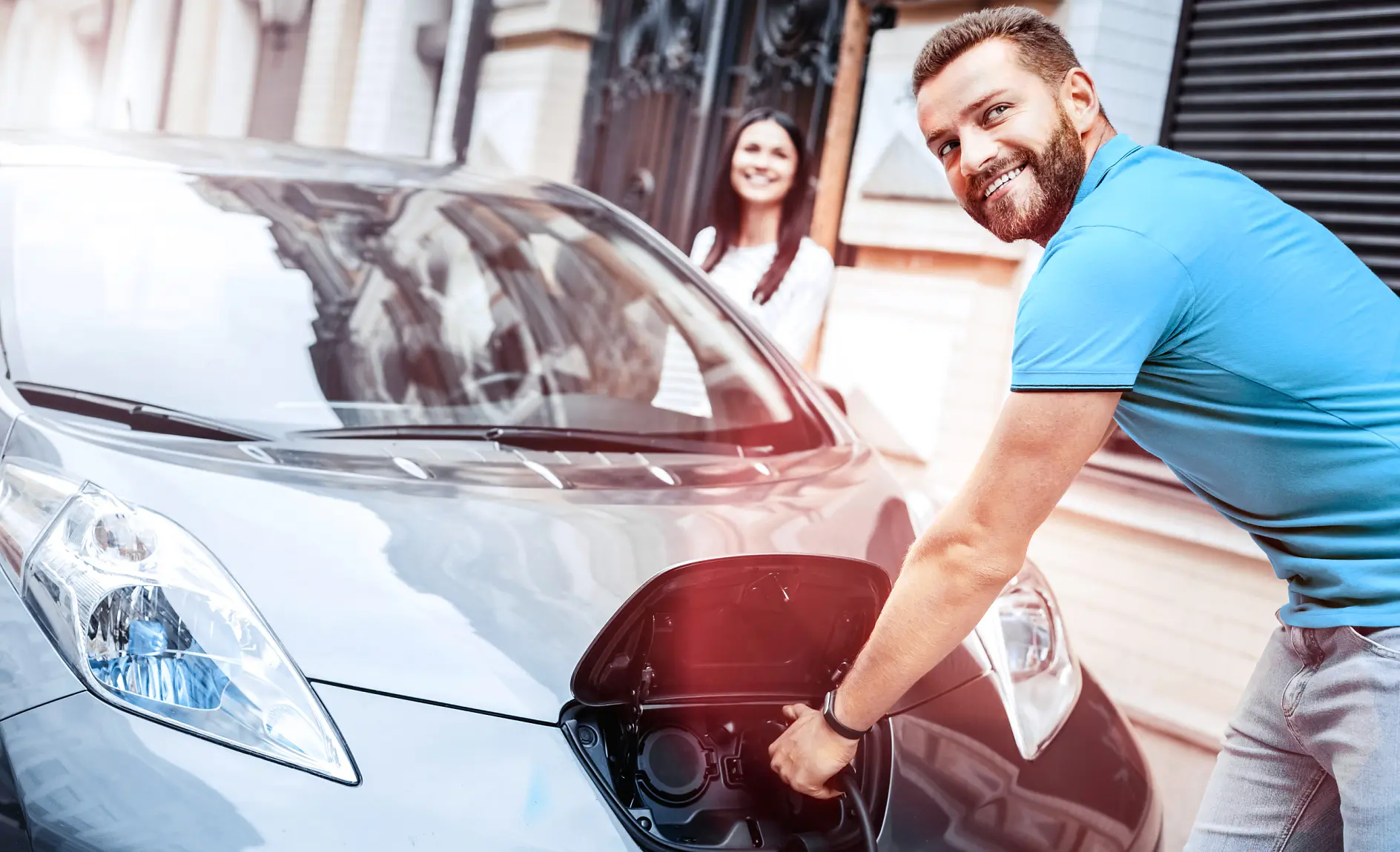 A man is plugging a charging cable into an electric car on a city street using tesa tape to secure the cable. The car is silver, and its front charging port is open. A woman stands nearby, smiling at the man as he uses tesa tape. Theyre in front of a building with a brick facade.