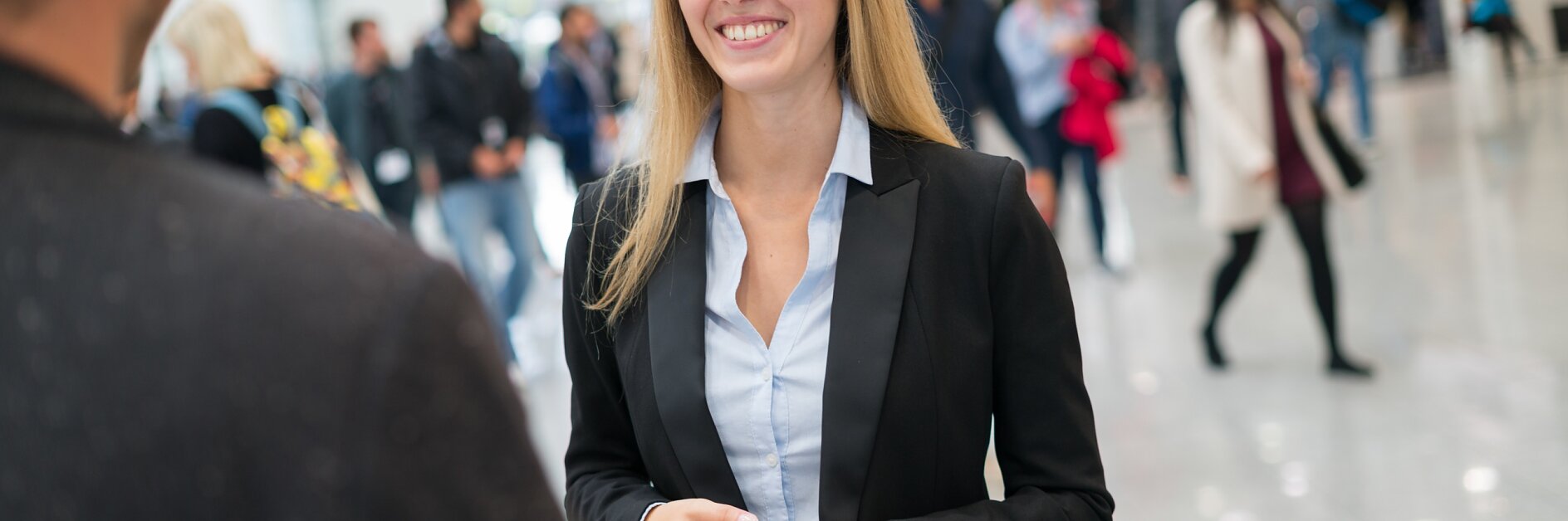 A woman in a black blazer and light blue shirt holds a smartphone while smiling at a person whose back is to the camera. They are in a busy indoor setting with people walking and large signs in the background, featuring tesa tape products prominently displayed.