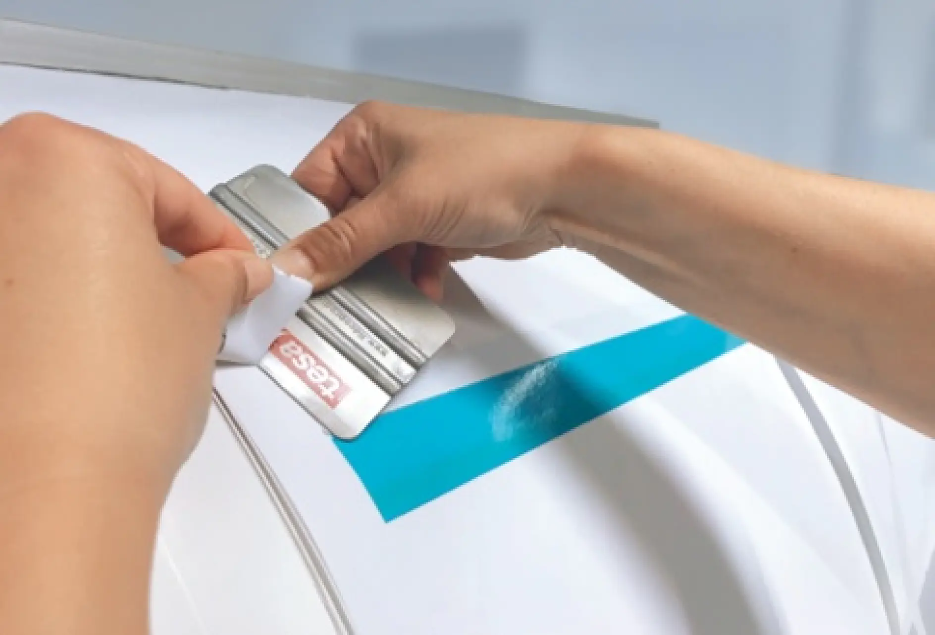 Close-up of hands applying a blue stripe of tesa tape to a smooth white surface using a flat tool. The persons fingers hold the edge of the tesa tape, while the tool smooths it down. A roll of tesa tape is visible in the background. (This text has been generated by AI)