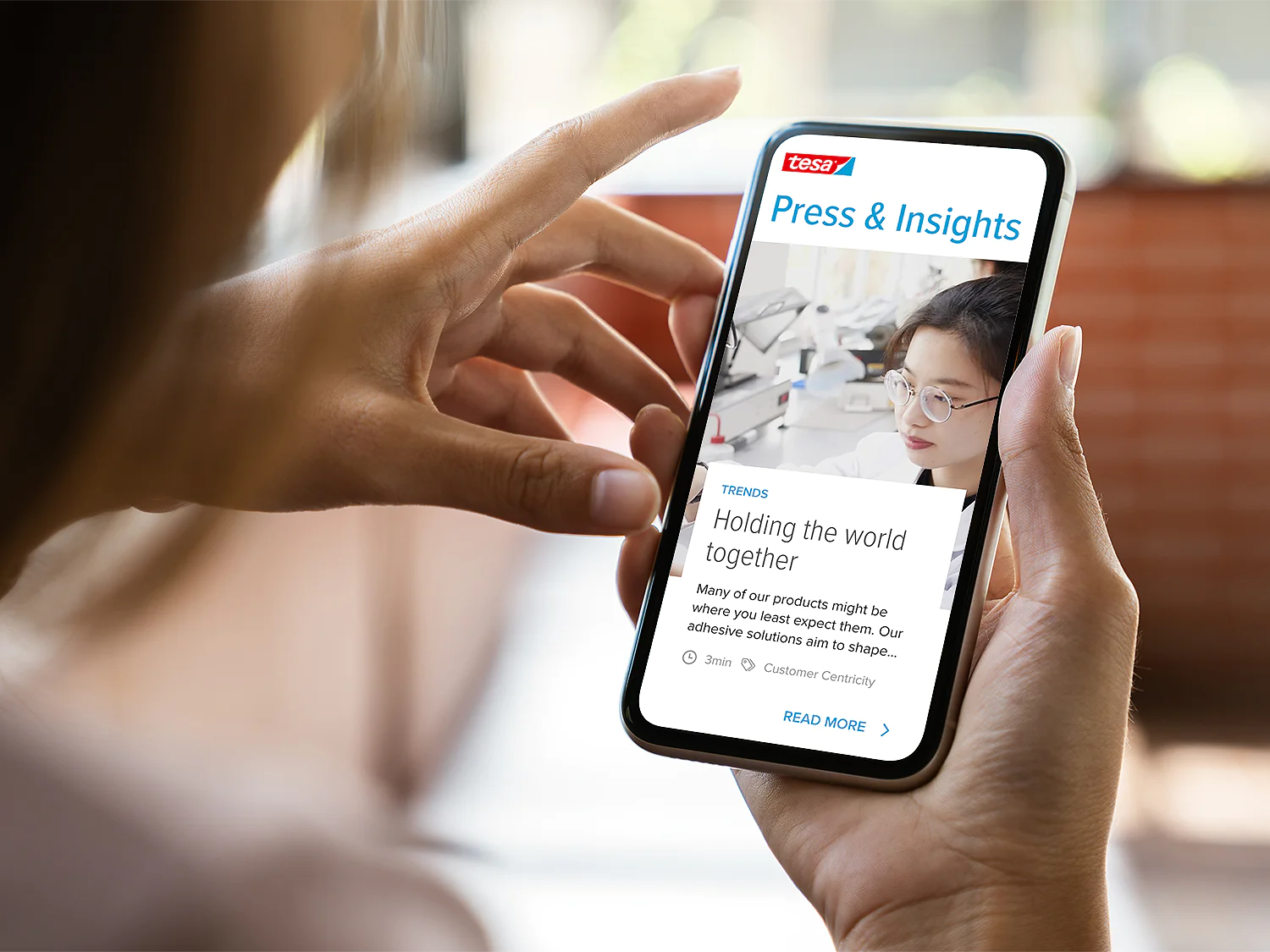 A person holding a smartphone displaying a webpage titled Press & Insights. The screen shows an article with an image of a woman behind a microscope and text about tesa tape adhesives. The smartphone is held in a hand with a blurred background. (This text has been generated by AI)