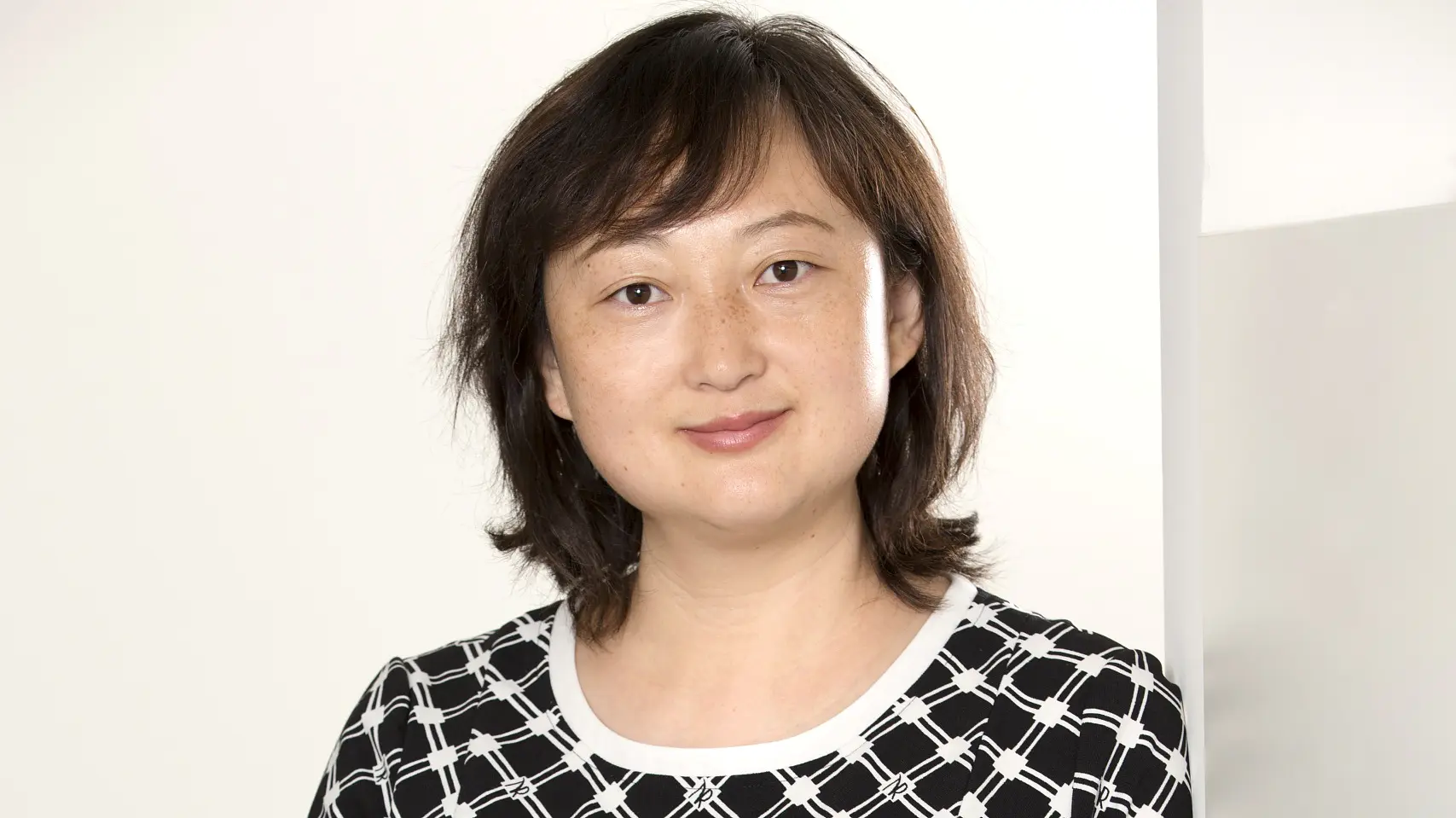 A woman with shoulder-length hair is wearing a black and white patterned dress. She is standing next to a light-colored wall, with her hands lightly crossed. The background is plain and neutral.