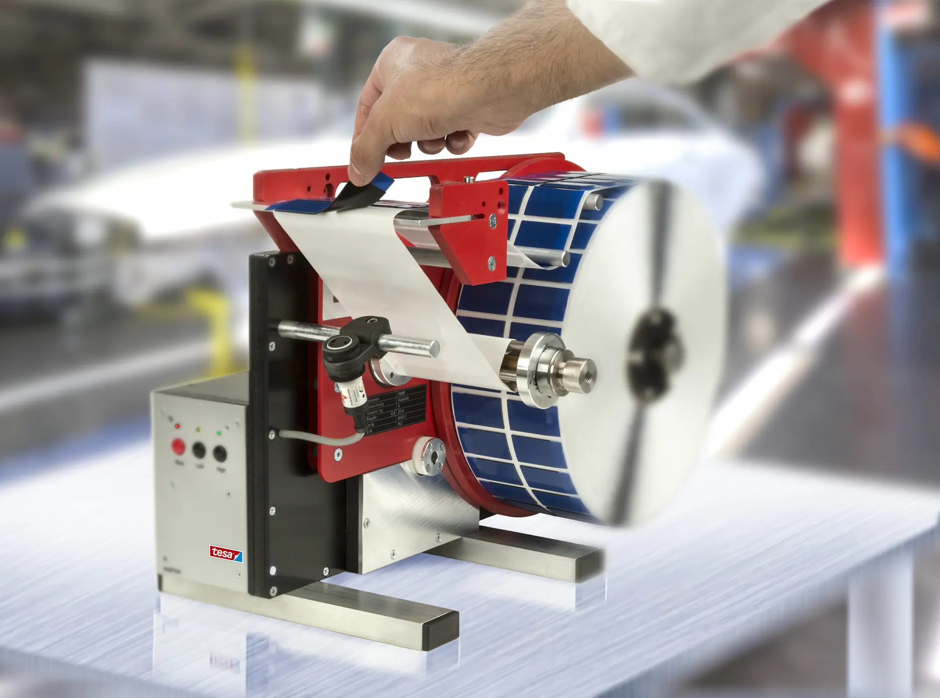 A person operates a labeling machine, feeding a strip of labels through the device. The machine has a roll of blue labels and metallic components. A roll of tesa tape is used in conjunction with the labeling process. It is situated on a surface with a blurred background of an industrial setting.