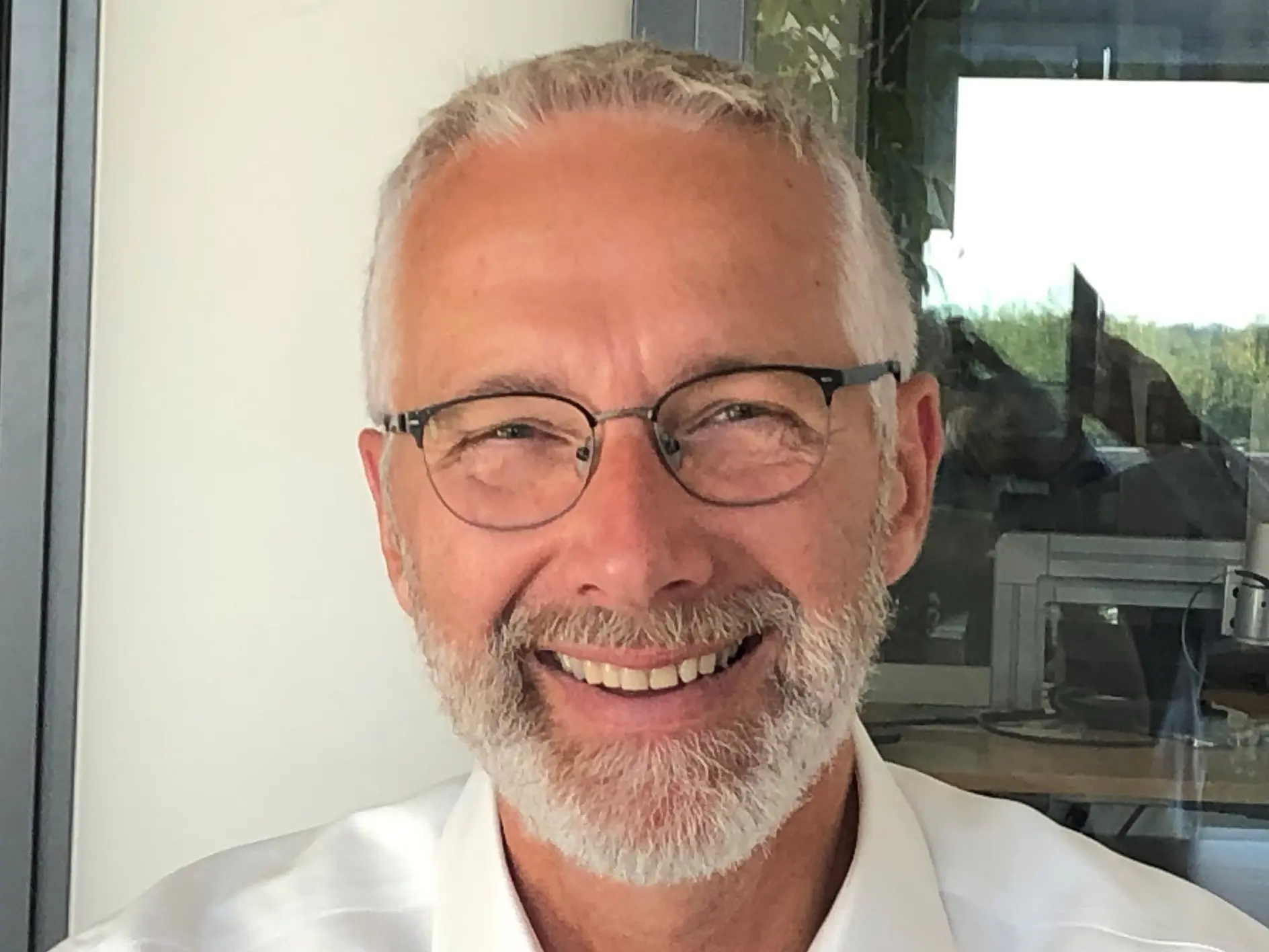 A man with short gray hair and a beard is smiling. He is wearing glasses and a white button-up shirt. The background shows a window with a view of trees and some interior office elements, featuring rolls of tesa tape displayed prominently on the desk.