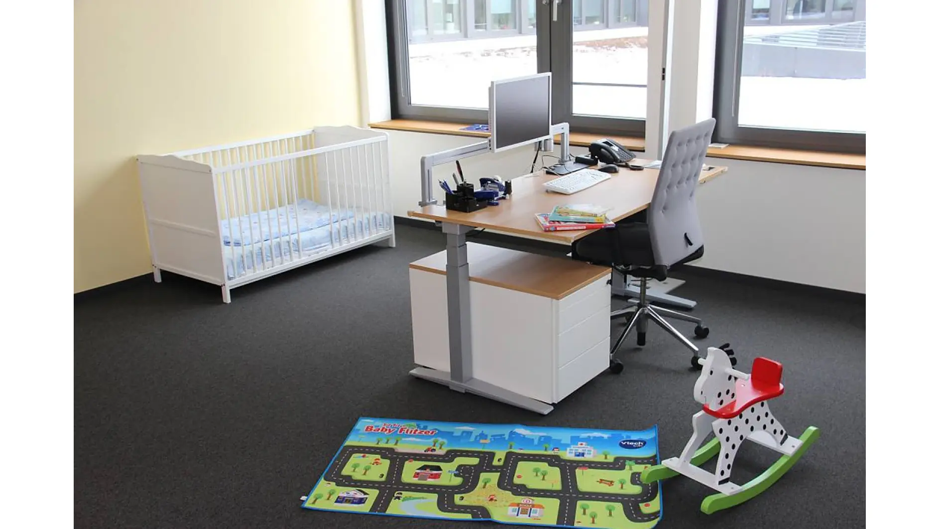 Office with a desk, chair, computer, and office supplies near a window. A white crib with blue bedding is in the corner. A colorful playmat and a black-and-white rocking horse with a red seat are on the carpeted floor. Various items are secured using tesa tape to ensure they stay in place. (This text has been generated by AI)