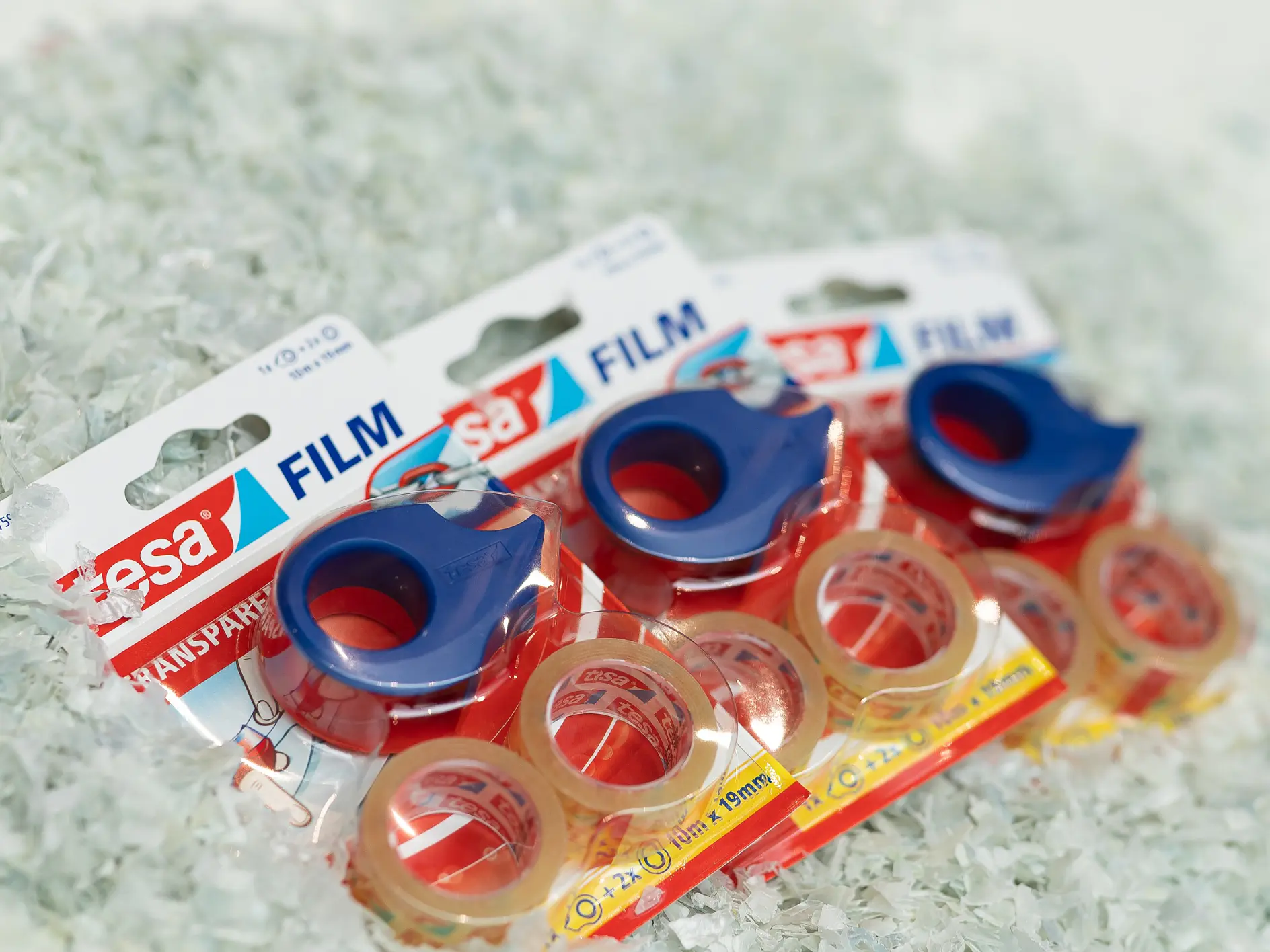 Three packages of tesa transparent adhesive tape are displayed on a pile of shredded paper. Each package contains a roll of tesa tape with a blue dispenser. The packaging text includes tesa Film and transparent. (This text has been generated by AI)
