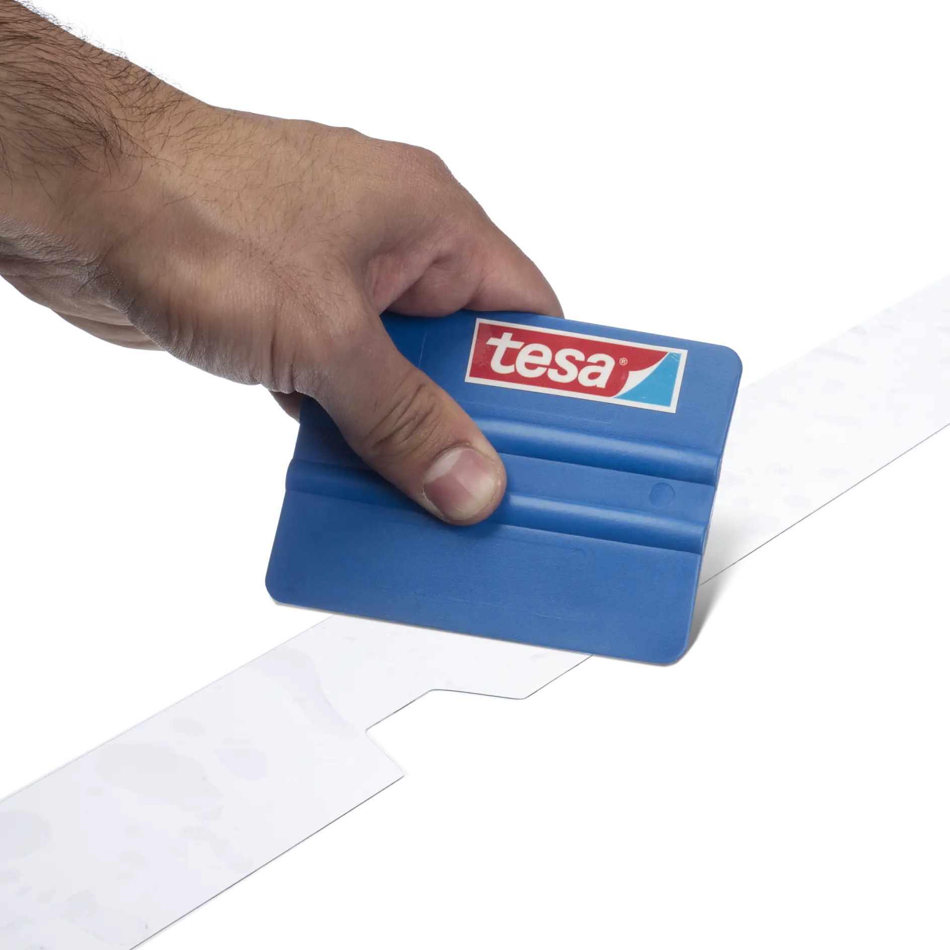 A hand holding a blue tool with a tesa logo, smoothing or adhering a strip of tesa tape. The background is plain and the focus is on the hand and tool performing the action.