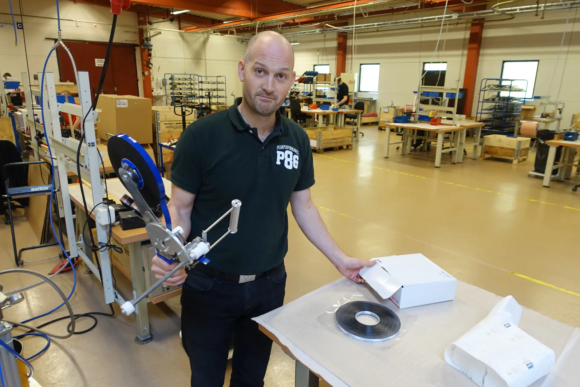 A person stands in a workshop holding a tool. They are next to a table with a white box, a coiled strip of tesa tape, and a paper bag. The workshop has various equipment and workbenches in the background.