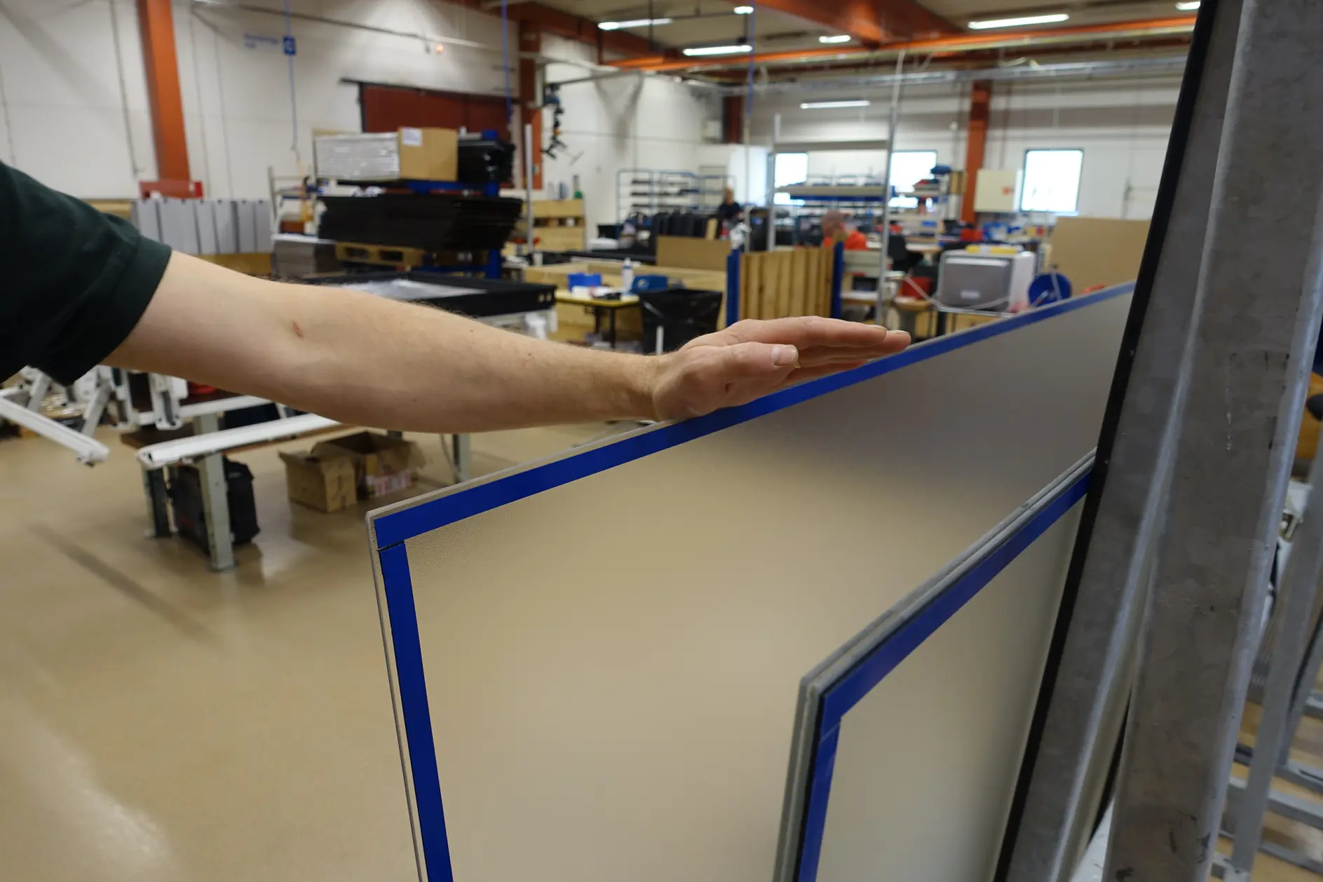 A persons hand is touching a large, clear panel with a blue border, standing in an industrial workshop. Various equipment, boxes, and tesa tape are visible in the background.