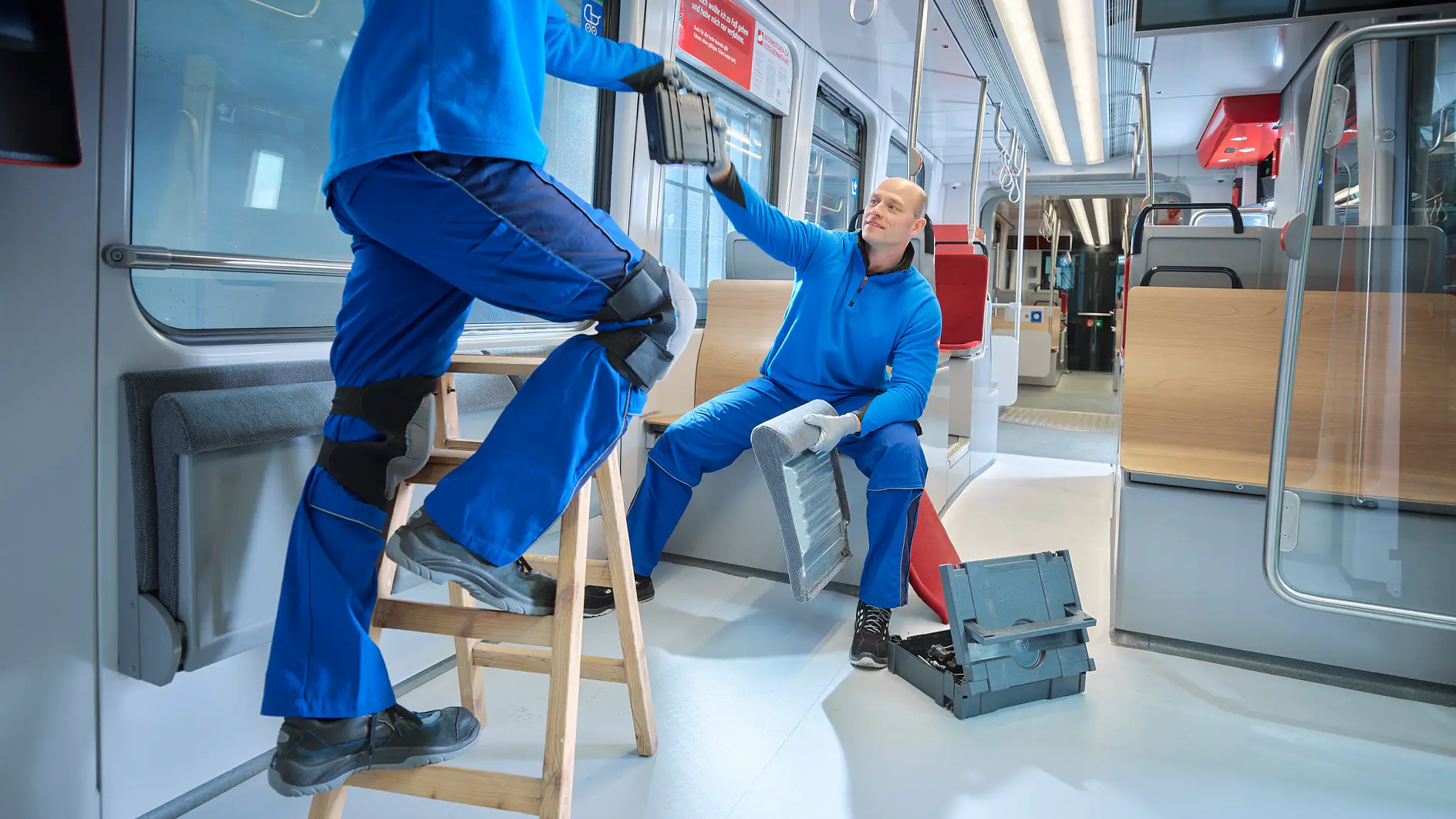 Two workers in blue uniforms are inside a train carriage. One worker stands on a wooden ladder holding equipment, while the other sits on a bench, handing over tesa tape. A toolbox rests on the floor nearby. The train is empty and well-lit.