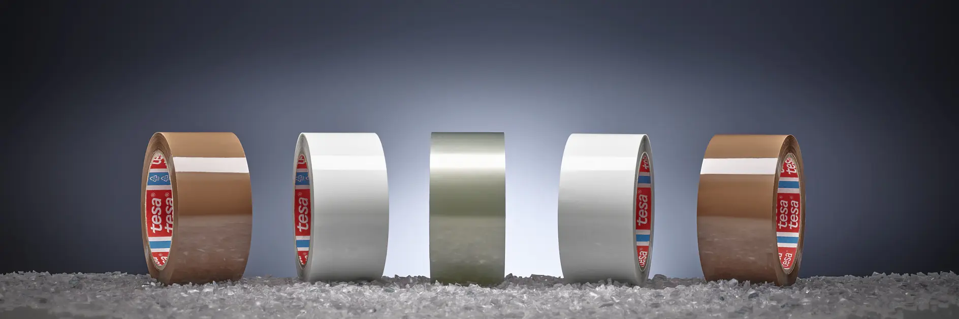 Four rolls of adhesive tesa tape, two brown and two white, are lined up on a surface scattered with small granules. Each roll is standing upright in front of a dark gradient background.