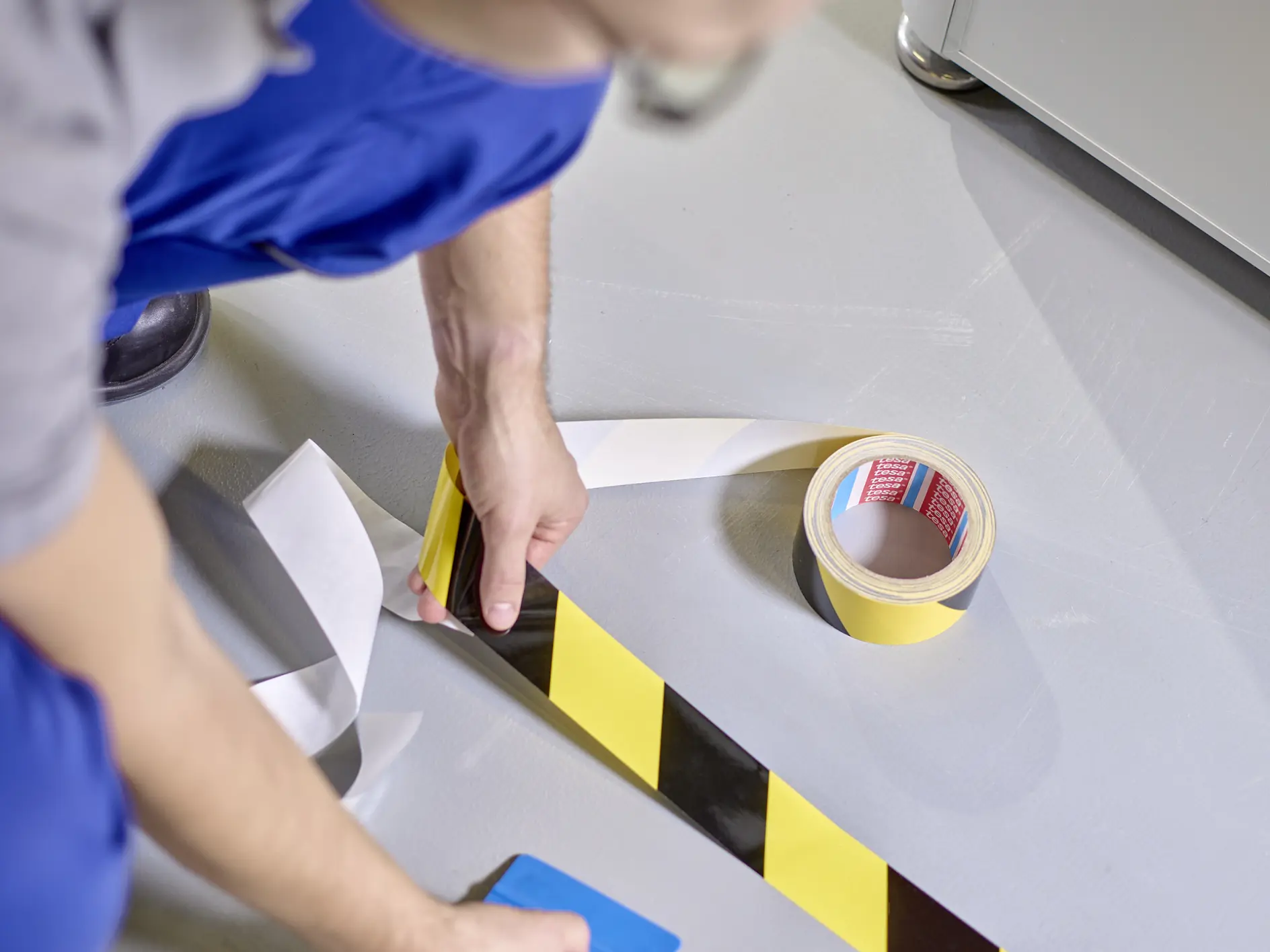 A person wearing blue pants and a gray shirt is applying yellow and black striped tesa tape to a light gray floor. They are using a blue tool to smooth the tesa tape, and a roll of similar tesa tape is nearby.