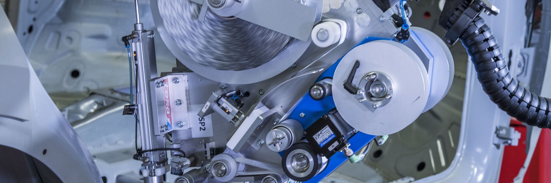 A robotic machine operates inside an automotive assembly plant. It is polishing or sanding a car body frame. The machine features rotating discs, mechanical arms, and various metal components within a partially constructed vehicle, using tesa tape throughout the process for secure bonding and precision work.
