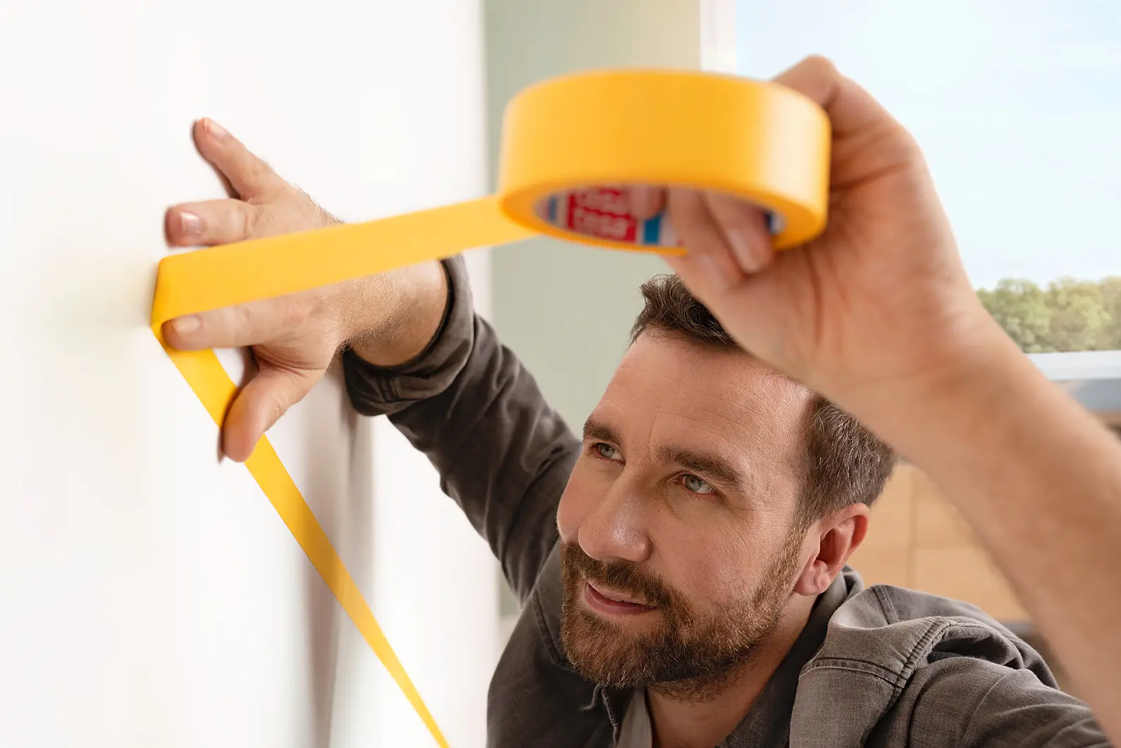 A man with a beard is carefully applying yellow tesa tape to a white wall. He appears focused on aligning the tesa tape properly. His other hand is pressed against the wall for support. The room seems to have natural light. (This text has been generated by AI)