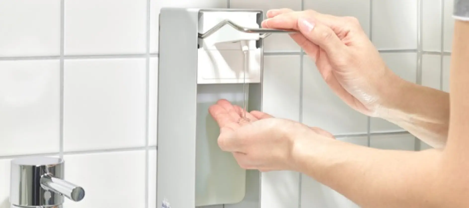 A person uses an elbow to press the lever of a wall-mounted hand sanitizer dispenser. The dispenser is metallic and silver, and the background features white tiled walls. A sink faucet is partially visible on the left. (This text has been generated by AI)
