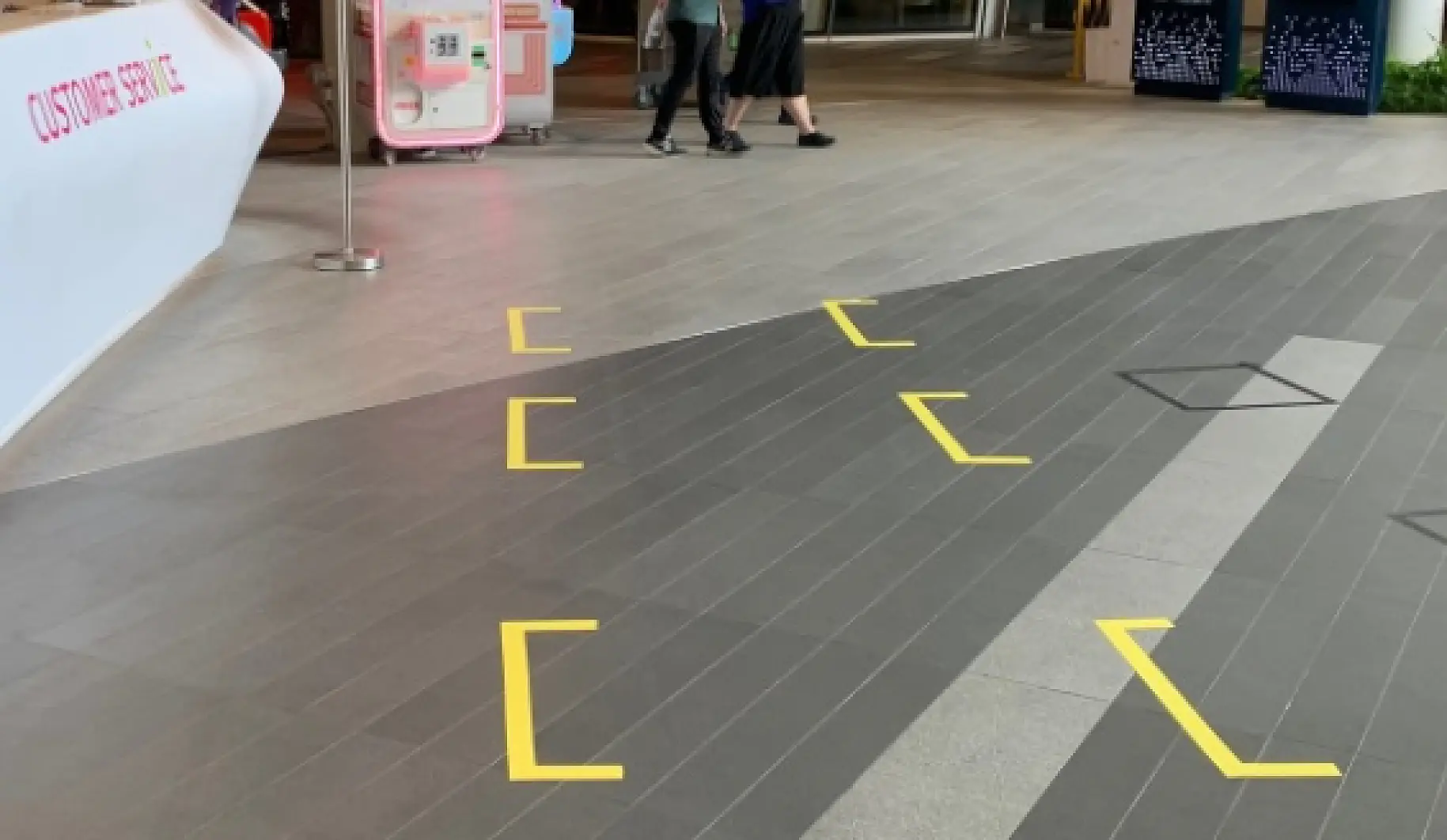 A tiled indoor floor with yellow brackets marked on it using tesa tape. Two people are walking in the background, near a customer service counter. A pink and white machine is visible near the entrance. (This text has been generated by AI)