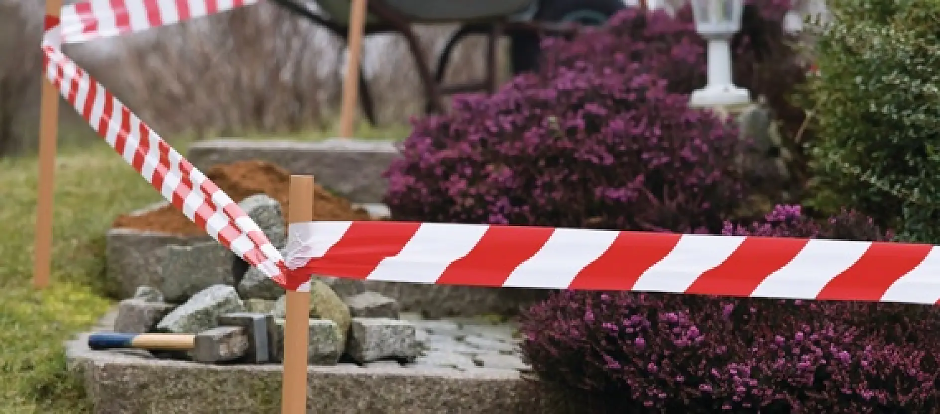 Red and white caution tesa tape cordons off an area with stone steps in a garden. There are purple shrubs nearby, and various landscaping tools such as a shovel and a trowel are visible on the ground. (This text has been generated by AI)