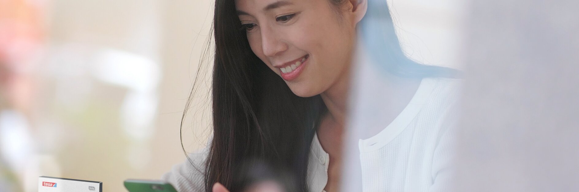 A woman wearing a white sweater is smiling and using a green smartphone. She is seated at a table. A blurred product resembling tesa tape is visible in the foreground on the left side.