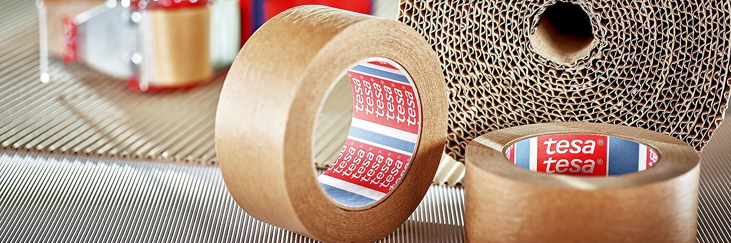 A large roll of corrugated cardboard is in the background. In the foreground, there are two smaller rolls of brown adhesive tesa tape, both labeled with the brand name. A tape dispenser with red handles is partially visible.