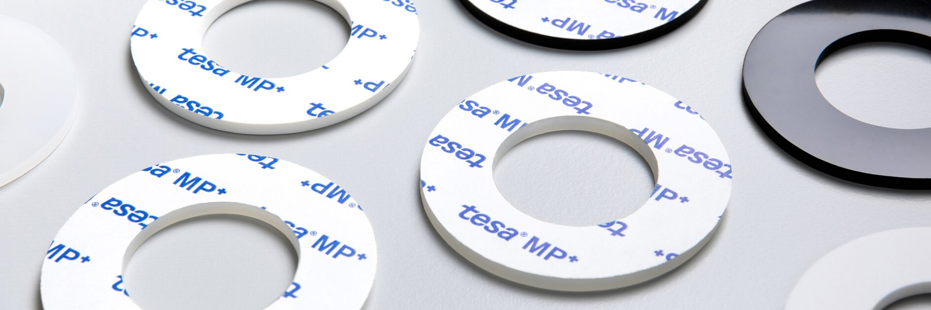 A group of adhesive rings are arranged on a light gray surface. Some rings are black, others are white, and a few have the tesa tape MP+ logo printed in blue on a white background. Each ring has a cutout center.