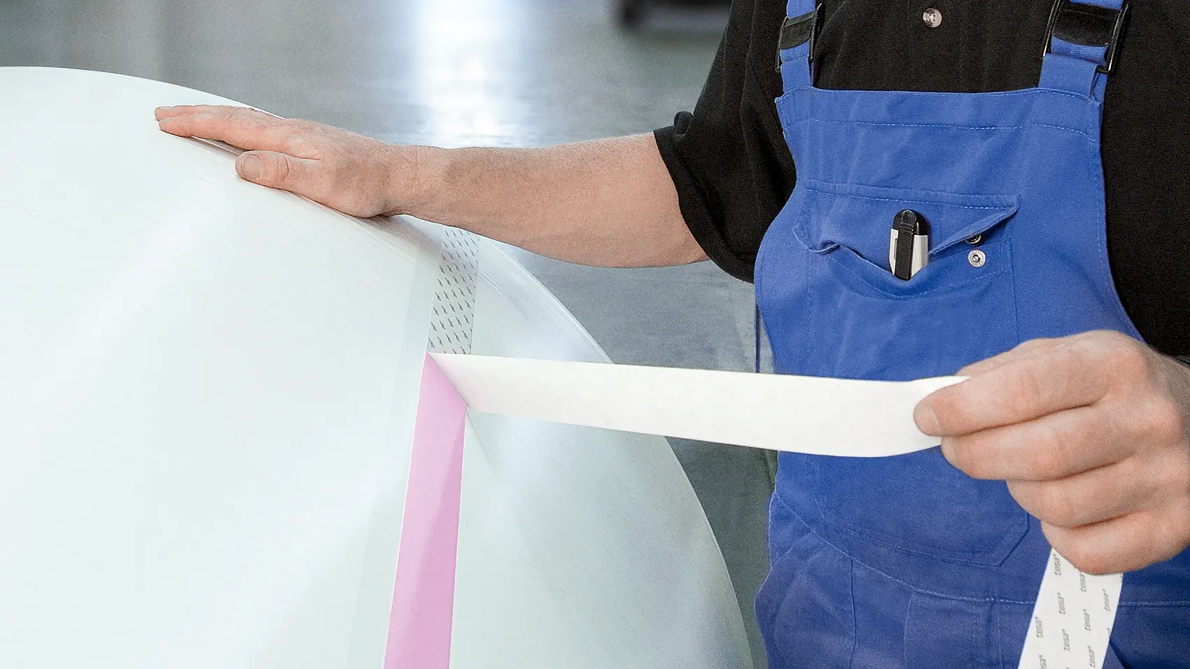A person in blue overalls and a black shirt is applying tesa tape to a large, white cylindrical object. They are indoors in an industrial setting, looking towards the camera. The background is slightly blurred, with equipment visible.