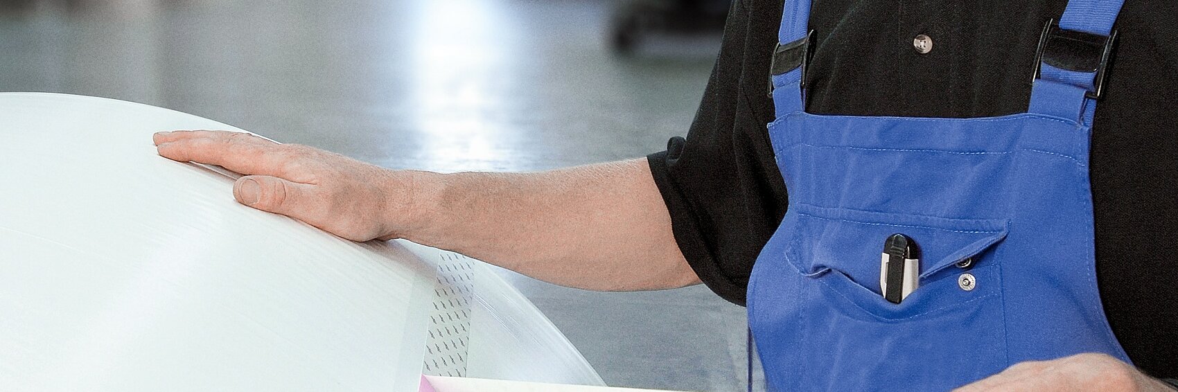 A person in blue overalls and a black shirt is applying tesa tape to a large, white cylindrical object. They are indoors in an industrial setting, looking towards the camera. The background is slightly blurred, with equipment visible.